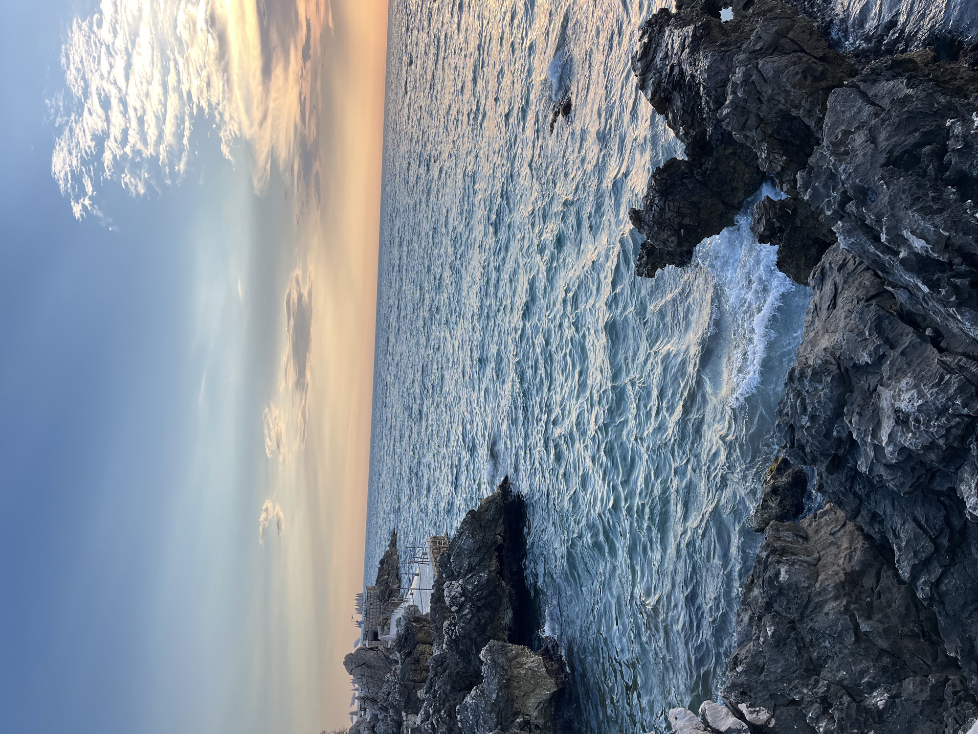 The image shows a rocky coastline on a clear day with a partly cloudy sky. The ocean is a deep blue with small waves lapping against the shore. In the distance, there is a walkway with a railing, and what appears to be beach umbrellas. The sky is a gradient of blue to orange, indicating either sunrise or sunset. The location is identified as Pečurice, Montenegro.