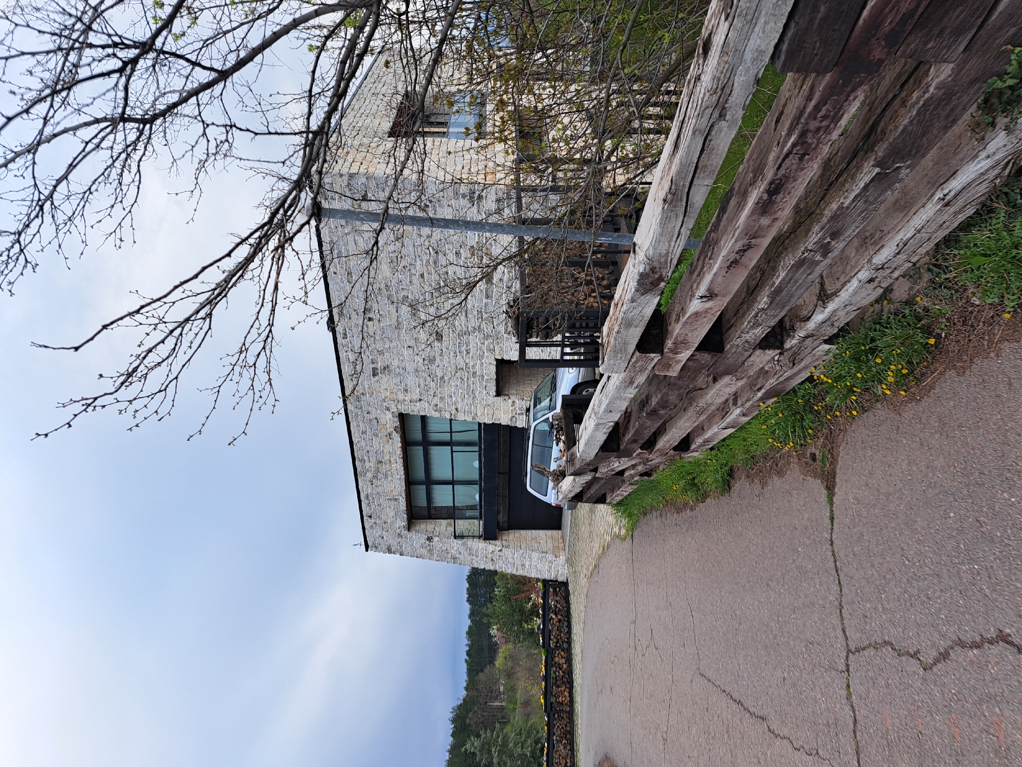 This image captures a tranquil rural or semi-rural scene in Lokorsko, Bulgaria, likely during early spring due to the bare tree branches with nascent buds. The dominant feature is a two-story building constructed from light-colored, neatly cut stone blocks, giving it a rustic yet sturdy appearance.
The building's facade features a small rectangular window on the upper floor. Below, a large, dark-framed window spans a significant portion of the ground floor, with a dark garage door positioned to its right. A horizontal metal beam or pipe is affixed to the building's exterior, partially obscured by tree branches. A light-colored SUV or van is parked in front of the building, partially hidden by the wooden fence and foliage.
In the foreground on the right, a robust, weathered wooden fence, possibly made from railway sleepers or thick timber logs, extends diagonally across the frame. Beyond the fence, green grass speckled with yellow dandelions grows along the edge of a paved asphalt road or path, which shows signs of cracking and runs from the bottom right towards the mid-ground.
The upper left portion of the image is dominated by a network of bare tree branches against a light, overcast sky, suggesting soft, diffuse daylight. Further back, beyond the stone building and the road, a neatly stacked pile of firewood is visible behind a dark railing, situated in front of a distant, verdant forested hillside under the same pale sky. No people or specific activities are visible, contributing to the quiet atmosphere of the scene. There is no visible text in the image.