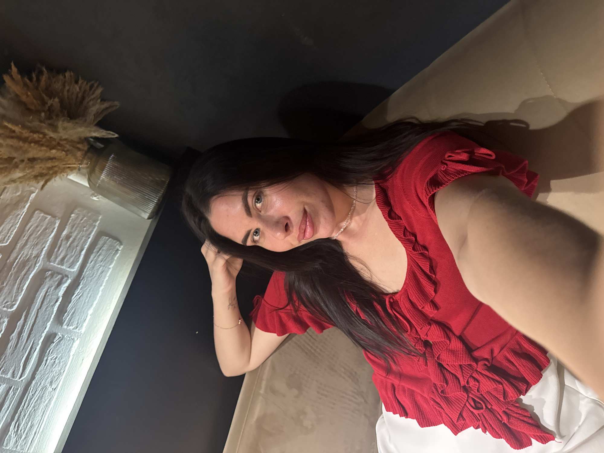 A woman with long, dark hair is lying on a sofa in a dimly lit room. She is wearing a red ruffled top and has her left arm behind her head. Her right arm is extended towards the camera, holding it for a selfie. The background includes a brick wall with a light fixture above it and a vase with dried reeds on a dark surface. The sofa appears to be upholstered in a light brown fabric. The image does not provide any information about the city of Chaikovsky, Russia.