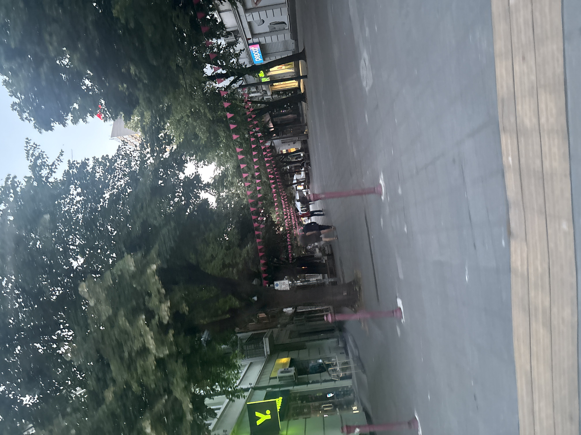 The image depicts a pedestrian street in Burgas, Bulgaria. On the left side of the street, there is a building with several shop fronts, one of which has a prominent bright green sign with a yellow "Y." logo. Further down the street, other shops are visible, including one with a "pepco" logo. A row of trees lines the street, and a string of pink triangular flags hangs across the walkway, suggesting a festive atmosphere or decoration. Several people are walking on the pedestrian area, appearing to be casual strollers. The ground is paved with grey tiles, and there are reddish-brown posts along the side of the walkway. The overall scene suggests a commercial and public space within the city.