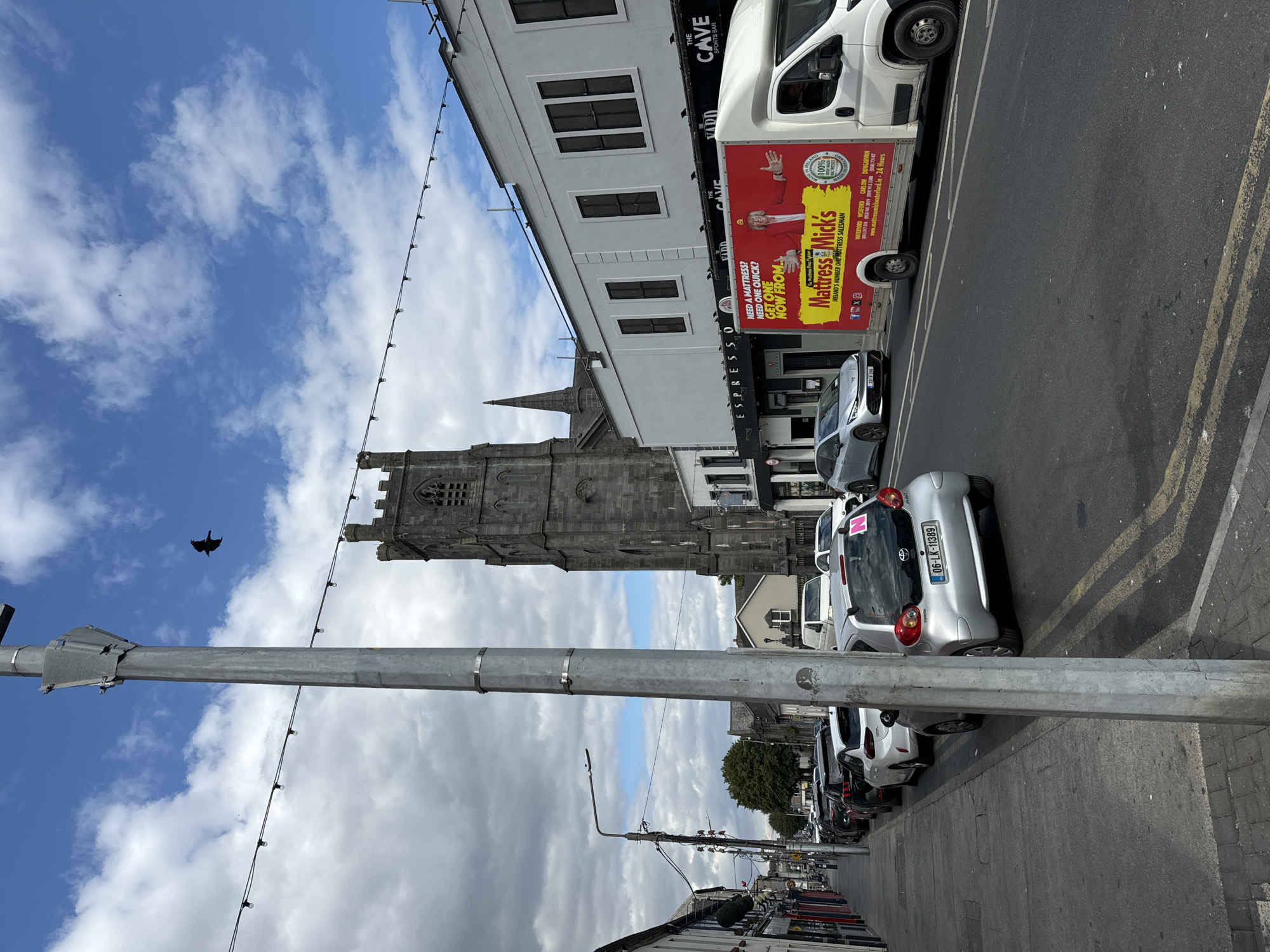 The photograph captures a street scene in Waterford, Ireland, with a prominent grey stone church tower dominating the background. In the foreground, a silver Toyota Aygo car is parked, displaying the license plate "06-LK-11389". To the right of the car, a white delivery truck is visible, featuring a large advertisement for "Mattress Mick's" with an image of a smiling man in a yellow hat. The truck also displays contact information and a website address: www.mattressmickwaterford.ie. The street is lined with parked cars and buildings, including businesses with signage such as "ESPRESSO" and "THE CAVE SPORTS BAR". A tall metal lamppost is positioned in the left foreground, and overhead, a string of lights spans across the street. A single bird is captured in flight against a blue sky with scattered white clouds.
