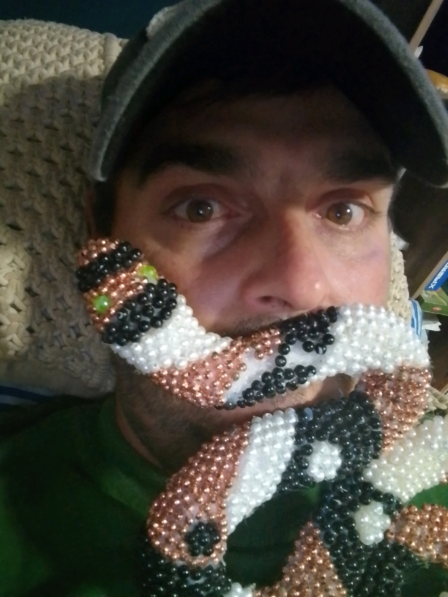 A man is taking a selfie. He is wearing a dark baseball cap and a green shirt. He has a beaded snake wrapped around his face, covering his mouth and part of his nose. The snake is made of white, black, and copper-colored beads, with two green beads forming eyes. The background appears to be a couch or chair with a textured cream-colored blanket. There is a box with some writing on it on the right side of the image. The man's eyes are open and looking directly at the camera. The lighting is artificial and casts shadows on his face. This image was taken indoors and does not provide any specific information about its location in the city of Wedel, Germany.