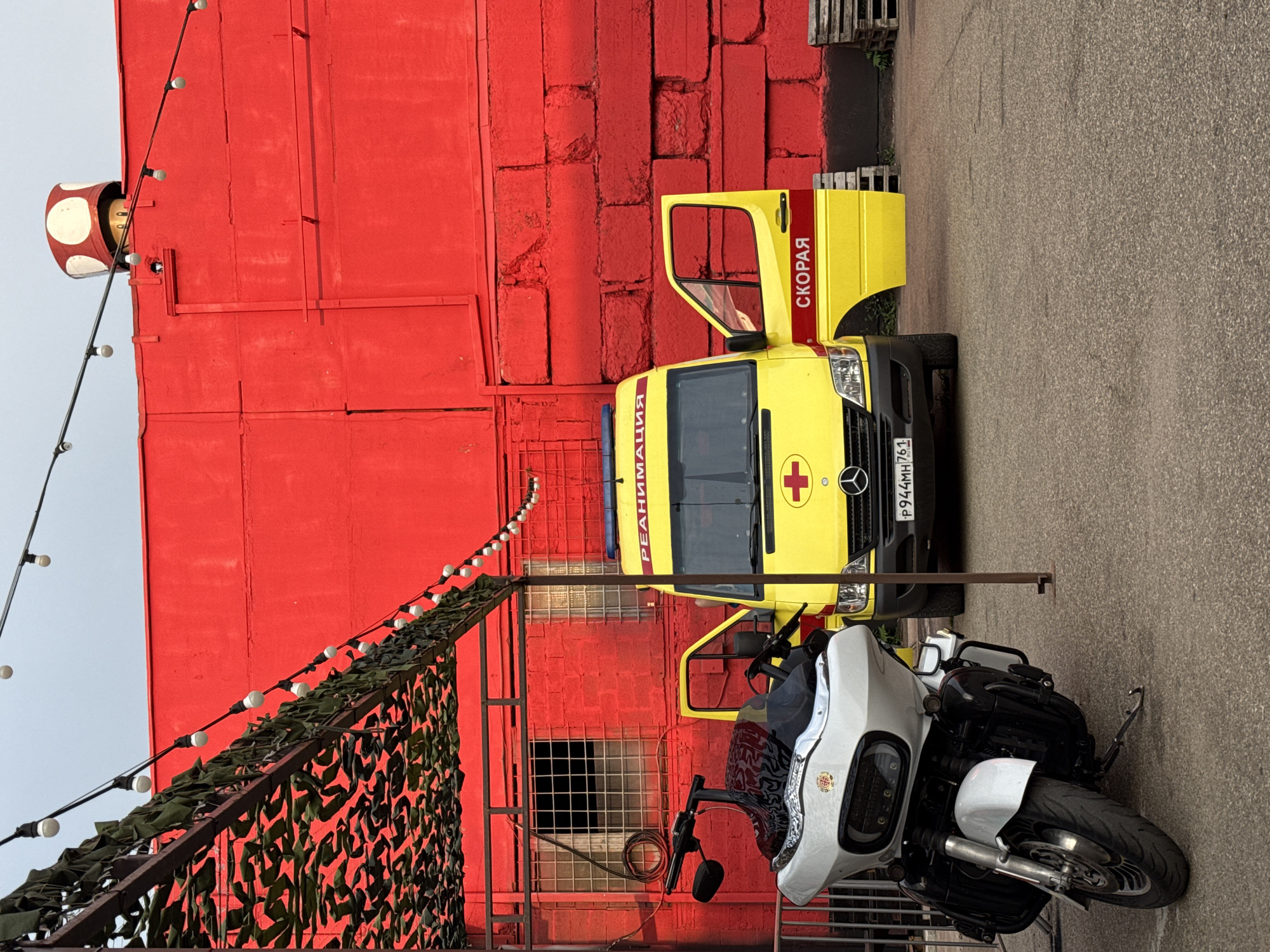 An ambulance, identified by the Cyrillic text "СКОРАЯ" (SKORAYA, meaning ambulance) and "РЕАНИМАЦИЯ" (REANIMATSIYA, meaning resuscitation), is parked in front of a vibrant red brick building. A white motorcycle is parked to the left of the ambulance. String lights with exposed bulbs are hung across the scene, some draped over a camouflage netting structure. The building has a rough, textured red surface and features a window with a metal grate. A mushroom-shaped object is visible on the roof of the building. The ground is paved asphalt. The license plate of the ambulance reads "P944MH 761 RUS".