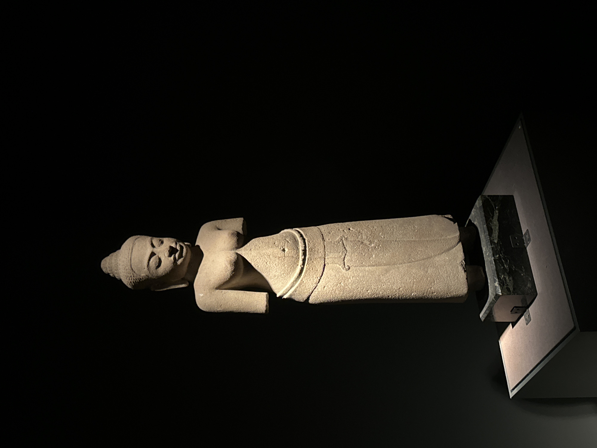 A stone statue of a female figure, possibly a deity or dancer, stands illuminated against a dark background. The statue depicts a woman with a headdress, exposed upper torso, and a long skirt wrapped around her lower body. She is standing on a dark, multi-faceted base, which is itself situated on a light-colored, angled display platform. The lighting highlights the texture of the stone and casts shadows, suggesting a museum or exhibition setting. This artifact is part of the collection at the National Museum of Korea in Seoul, South Korea.