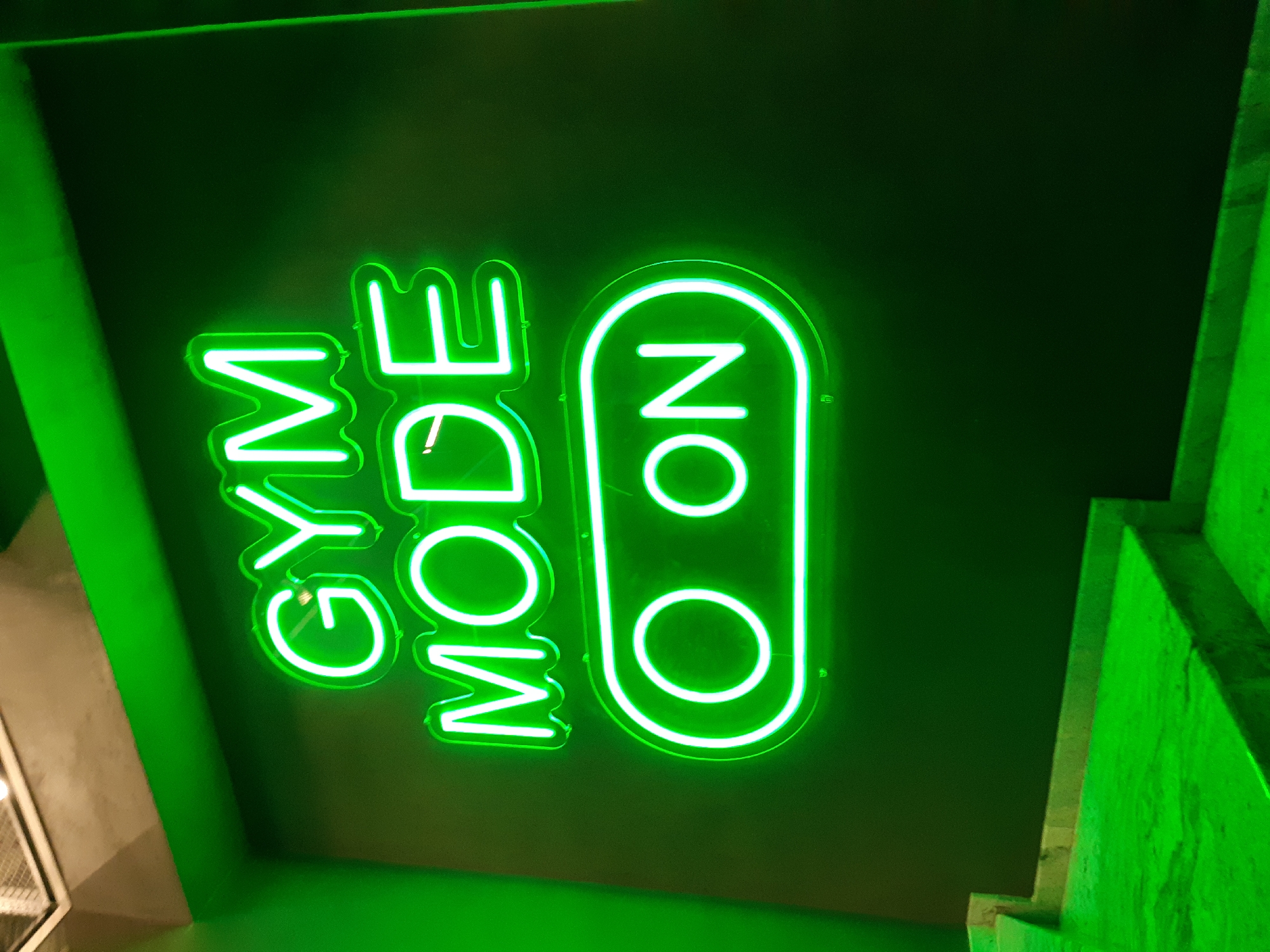 A photograph shows a neon green sign that reads "GYM MODE ON". The sign is mounted on a dark green surface. The photo appears to be taken indoors, and there is a bright green wall corner visible in the lower-right. The picture does not contain information about any people or Plovdiv location.