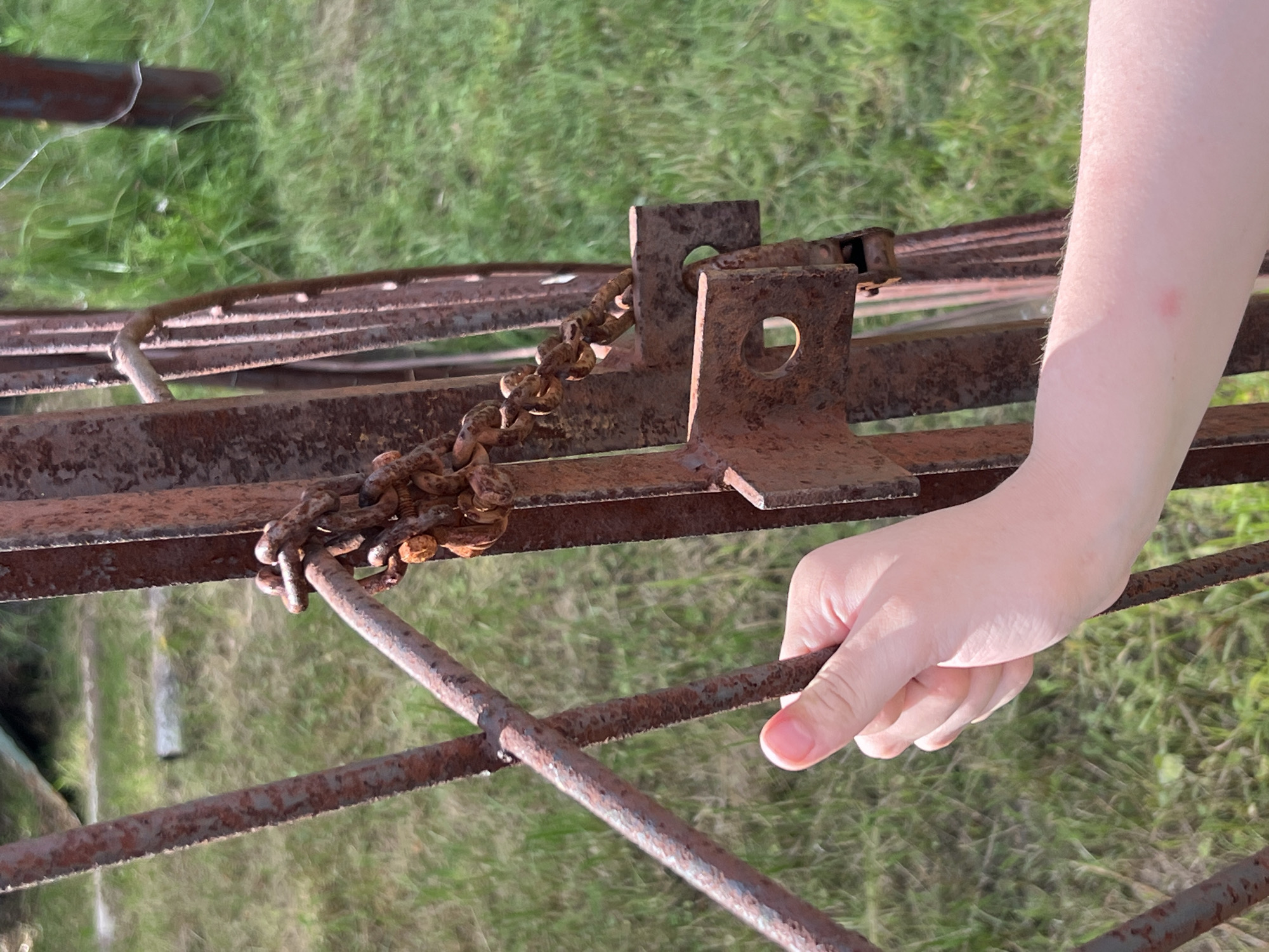 A hand is holding onto a rusted metal gate. The gate is composed of vertical bars and has a chain wrapped around one of the vertical bars, securing it. The chain appears old and also rusted. In the background, green grass is visible, suggesting an outdoor setting. The image is taken from a close-up perspective, focusing on the hand and the gate. The specific location is Strakhovo (Sitnikovskii S/S), Russia.