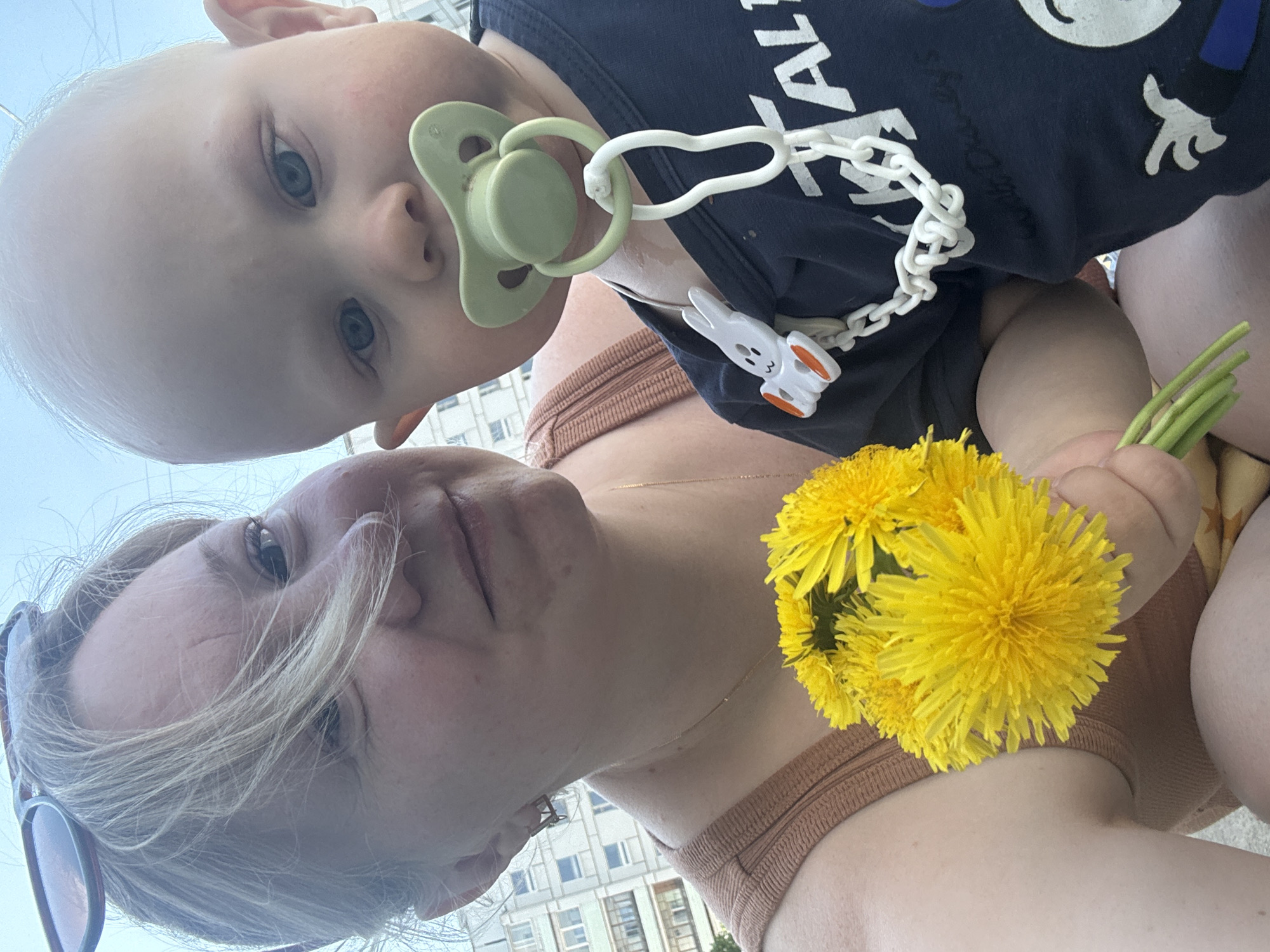 A selfie taken by a light-skinned woman and a baby. The woman is looking up at the camera, and is wearing a tan-colored top, a necklace, and her sunglasses are tucked into the top of her head. She is holding a bouquet of yellow dandelions. The baby, with fair skin and blue eyes, is above her, sucking on a pacifier. The pacifier is attached to a white chain, and the chain is clipped onto the baby's dark blue shirt. The background shows a building, suggesting an urban environment. The image was taken in Chaikovsky, Russia.