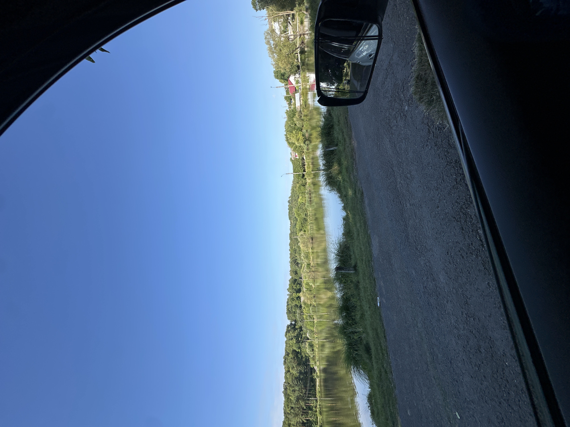 The image depicts a view from inside a vehicle, looking out towards a tranquil body of water lined with lush green trees and vegetation. The sky is a clear, bright blue, suggesting a sunny day. In the distance, several houses with red roofs are visible, nestled amongst the trees, indicative of a rural or semi-rural setting. A side-view mirror of the vehicle is visible in the foreground, reflecting the car's interior and a portion of the surrounding landscape. The foreground also shows a gravel road adjacent to the grassy bank of the water. The scene appears to be a calm and scenic landscape, likely captured during daylight hours. The location is identified as Makiv, Ukraine.