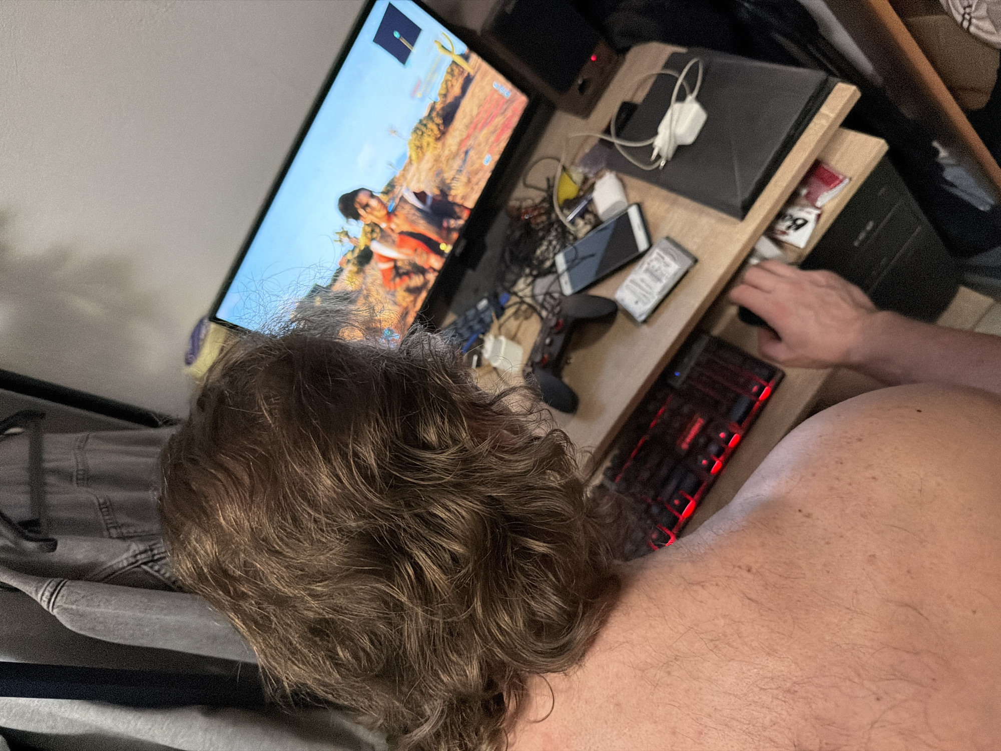 A person is seated in front of a computer setup, focusing on the screen displaying a video game. The desk holds a keyboard, mouse, mobile phone, external hard drive, and a computer tower. Assorted cables and electronics are scattered around the desk. The person has brown curly hair. The environment includes a bed, clothing, and storage space in the background. There is no visual information to suggest the location is Saint Petersburg, Russia.