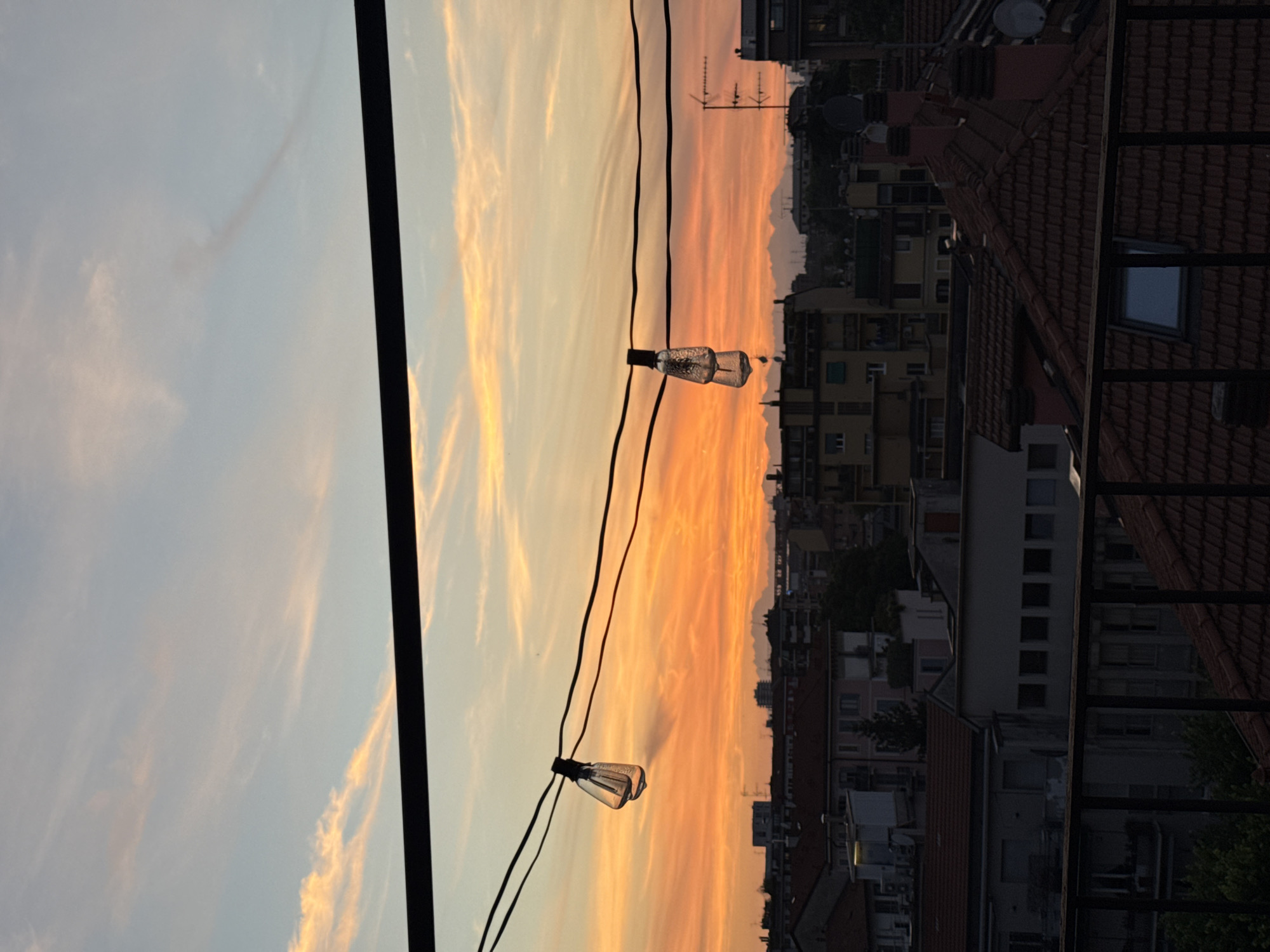 The image shows a sunset view over Milan, Italy, framed by a balcony. The foreground features two string lights suspended from a cable. Behind the lights, the sky displays a gradient of colors, transitioning from pale blue to orange. In the distance, a dense urban landscape unfolds with rooftops, buildings, and antennae visible. The city skyline is silhouetted against the bright sky. A black frame appears on the left side of the image, likely part of the balcony structure.