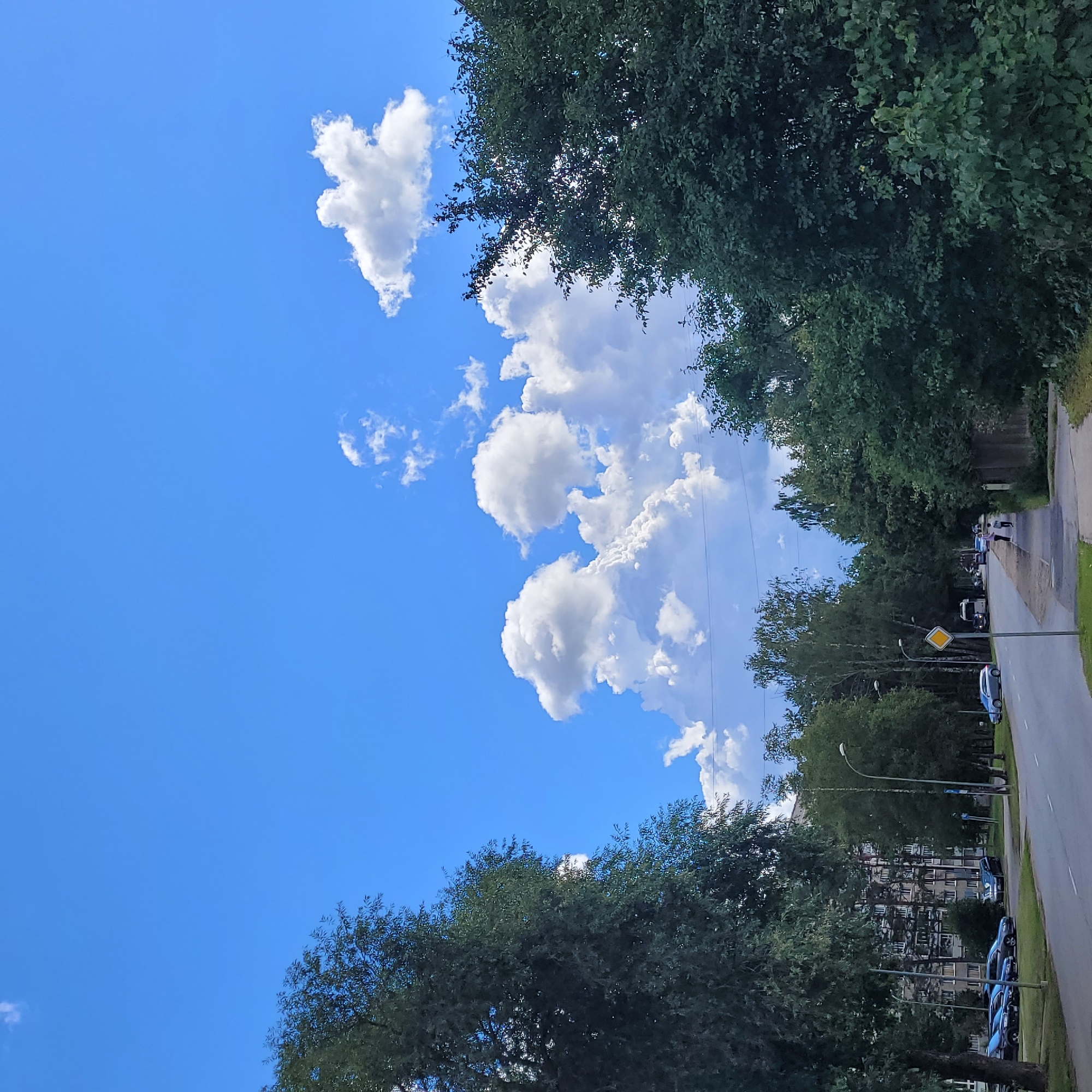 The image depicts a street scene in Jūrmala, Latvia, on a sunny day with a bright blue sky dotted with fluffy white clouds. A paved road runs diagonally from the bottom left to the center right of the frame. Cars are parked along the left side of the road. On the left side of the road, a tall, dense tree with dark green foliage dominates the foreground. Further down the street on the left, a multi-story apartment building with many windows is visible behind a line of trees.
The right side of the road features a sidewalk, a wooden fence, and more lush green trees and bushes. A street lamp is present on the right side of the road. Two figures, appearing to be a child and an adult, are walking on the sidewalk in the mid-ground. In the background, more trees and buildings can be seen. The overall impression is a suburban or residential area with a tree-lined street.