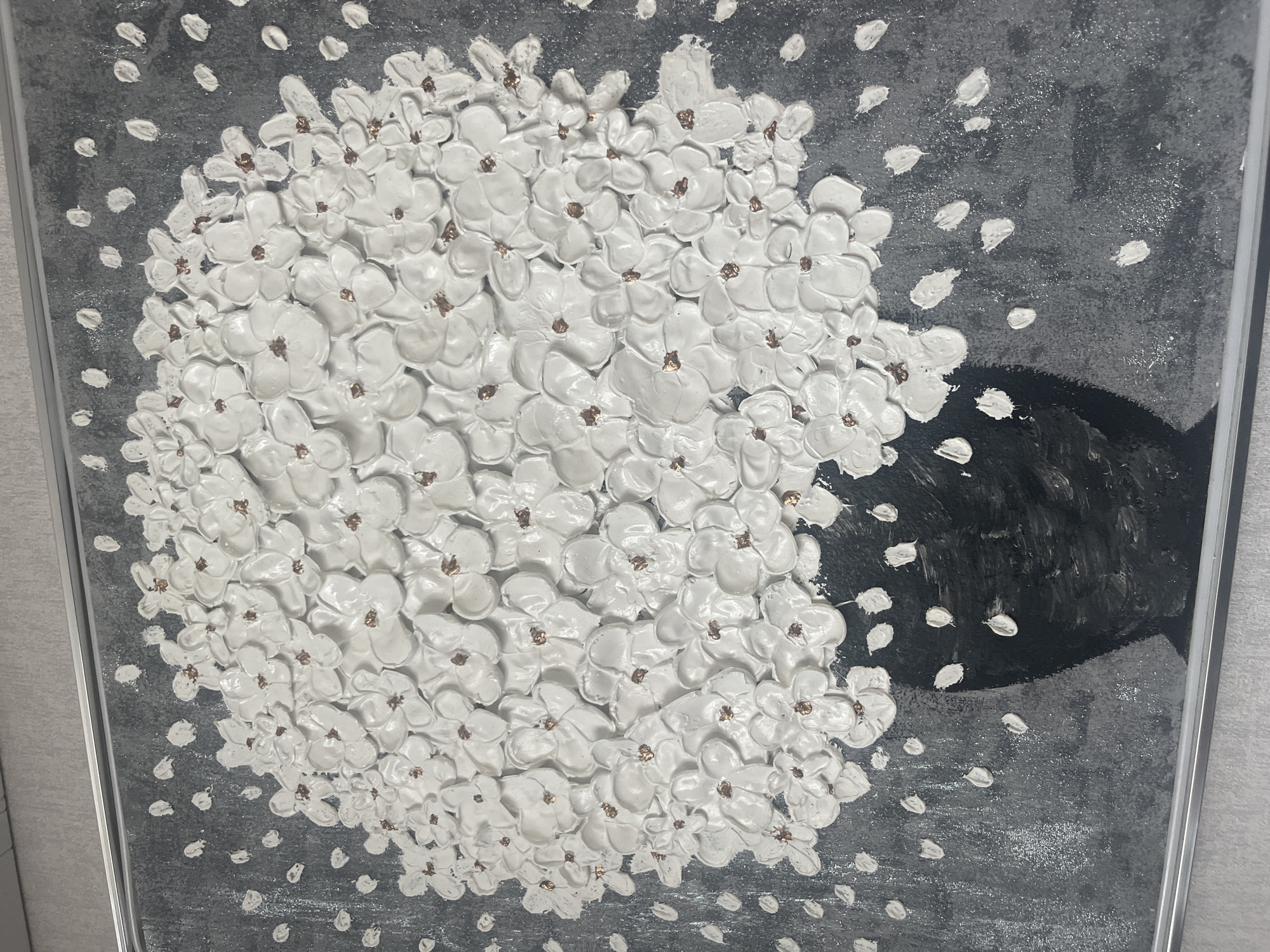 The image is a close-up of a framed textured artwork, likely a painting or a mixed-media piece. The artwork depicts a large cluster of white, three-dimensional flowers with small, coppery-colored centers, arranged in a rounded bouquet. The flowers are densely packed and appear to be made of a thick impasto material. They are set against a dark, textured background with a greyish hue. Scattered around the bouquet, and seemingly falling from it, are numerous small, white, petal-like or dot-like elements. At the bottom of the bouquet, a dark, abstract shape suggests a vase or pot. The overall style is abstract and textural. The question about the location in the city of Tyumen, Russia, cannot be answered as the image itself provides no such information.