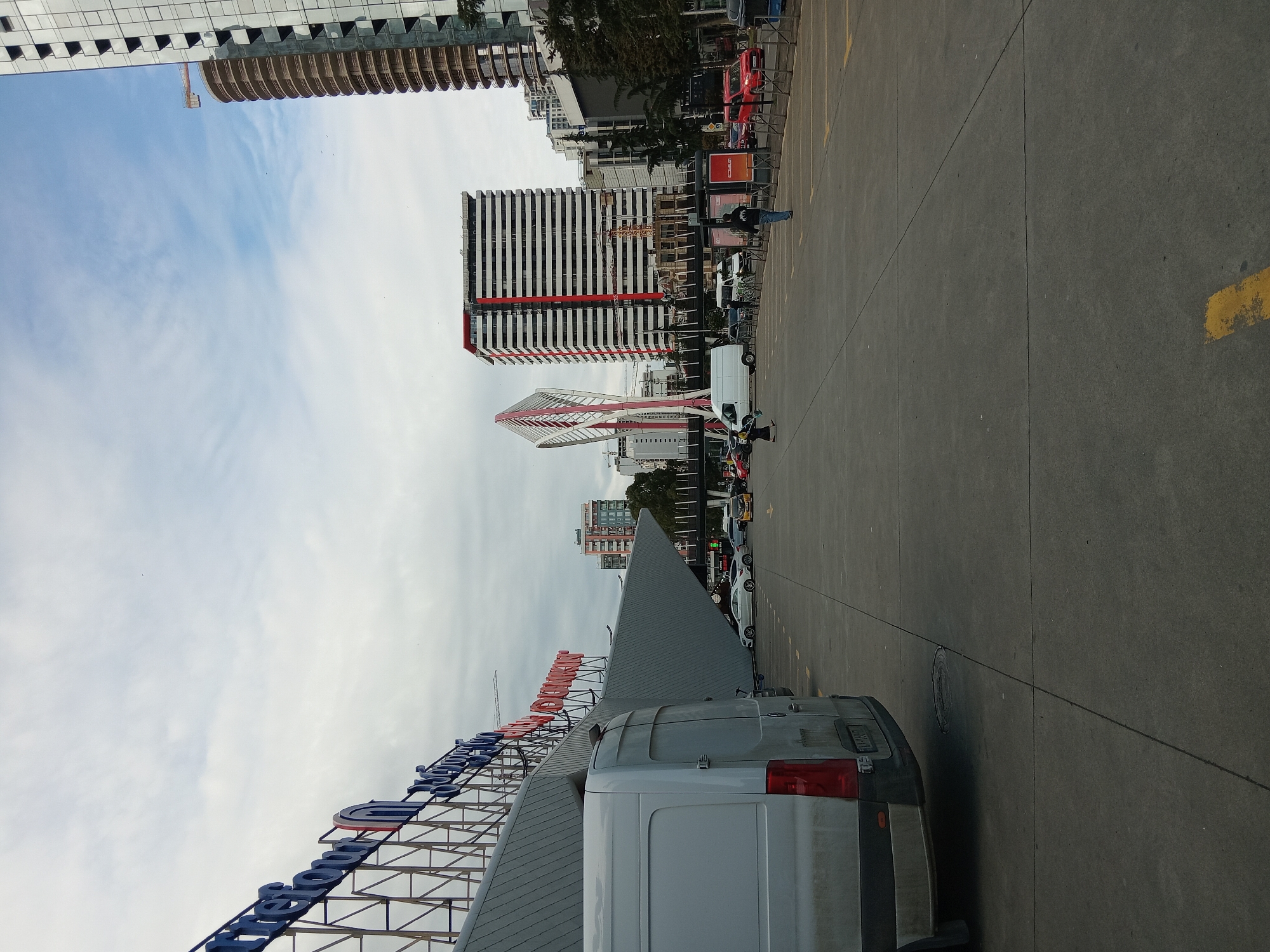 The image shows a view of the city of Batumi, Georgia, looking towards the skyline from a parking area adjacent to what appears to be the Batumi Grand Mall, indicated by a large sign. The sky is overcast with clouds. Several modern buildings dominate the skyline, including a distinctive red and white structure (likely Radisson Blu Hotel), a striped white and red building under construction, and a tall skyscraper further down the street. Cars and vans are parked in the parking area. There are people walking in the area adjacent to the parked cars. The perspective suggests the image was taken from a relatively low vantage point, possibly from within the parking lot or at its edge. A white van is parked in the foreground. The road in the parking area has yellow parking stripes.