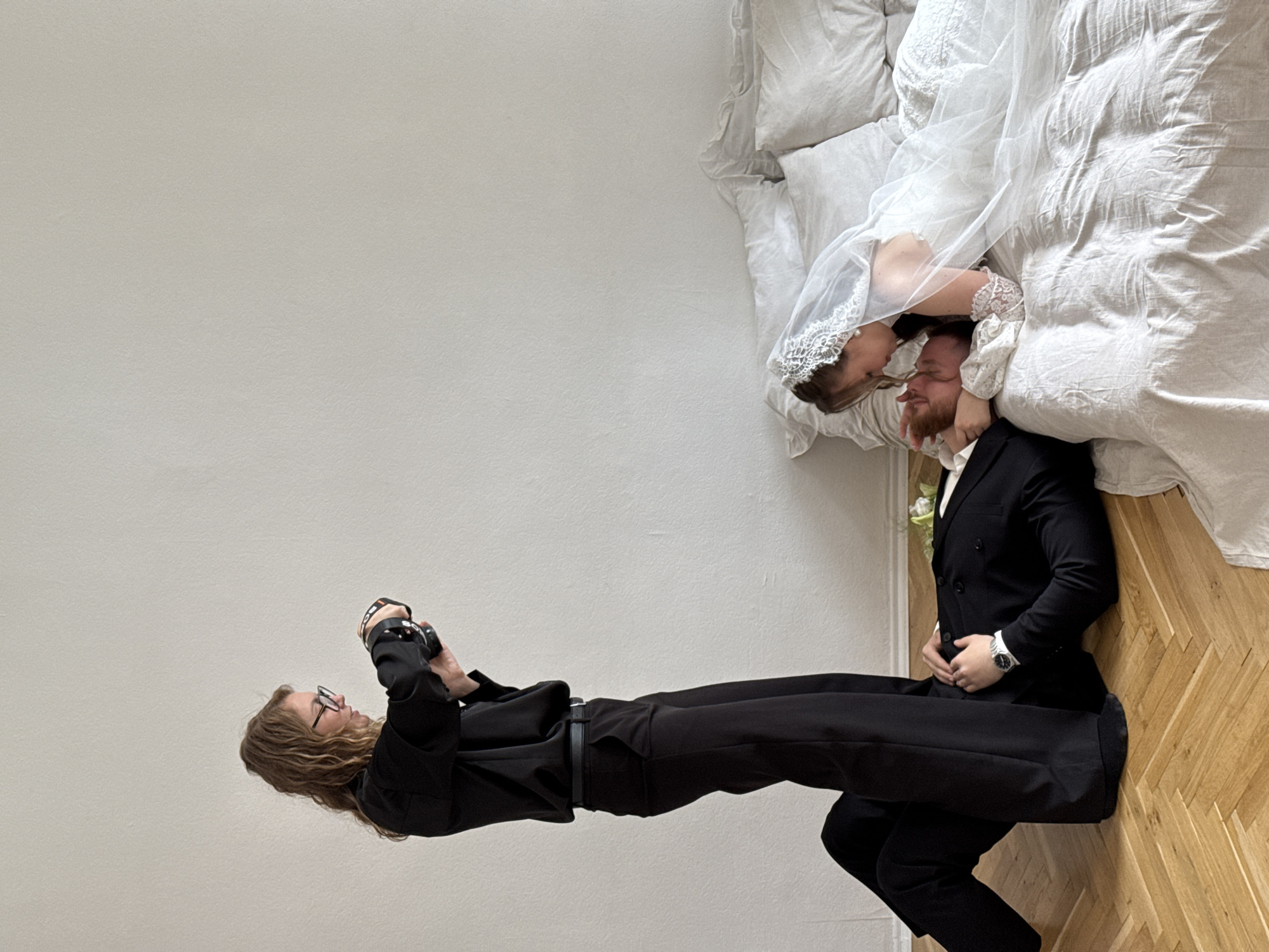 The scene depicts an indoor wedding photoshoot in Perm, Russia, featuring three individuals and a bedroom setting.
On the left, a woman identified as a photographer stands over a man. She has wavy, light brown hair and wears dark-rimmed glasses, a black long-sleeved shirt, black trousers, and a black belt. Her left leg is bent, with her foot resting on the man's mid-torso, while her right leg is straight. She holds a black camera with both hands, looking down at its screen or controls, suggesting she is adjusting settings or reviewing an image.
On the wooden parquet floor below the photographer, a man dressed in a black suit with a white shirt lies on his back. He has short reddish-brown hair and a beard, and a silver watch is visible on his left wrist. His arms are bent, with his hands resting on his chest or stomach. He gazes upwards towards the woman on the bed. A small cluster of white and green flowers lies on the floor near his head, close to the bed.
To the right, on a bed with crumpled white linen sheets and multiple pillows, a woman dressed as a bride is positioned. She wears a white wedding dress, a lace-edged veil over her head and hair, and white lace sleeves or gloves. She leans over the man on the floor, her face close to his, suggesting an intimate moment or a kiss. Her left hand is near his head/neck area. Her body extends along the bed, which occupies the right half of the image.
The background consists of a plain, light off-white or beige textured wall. The floor is made of light brown wood with a chevron parquet pattern. The overall environment is minimalist and brightly lit.