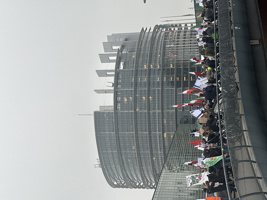 The image displays an outdoor scene in Strasbourg, France, featuring the European Parliament's Louise Weiss building under an overcast grey sky. The building, a large, modern structure with a curved glass facade and distinctive rectangular architectural elements on its roof, dominates the background. In the midground, a crowd of approximately 50-100 individuals is gathered on a pedestrian bridge or walkway.
The people are actively participating in a demonstration, holding numerous flags and rectangular signs. A significant number of flags are vertically-striped green, white, and red, many featuring a central emblem such as a stylized lion and sun. Several blue and white flags displaying a Star of David (Israeli flags) are also visible, interspersed with solid red and green flags. Participants are dressed in various cold-weather attire, including coats and hats. Their collective orientation suggests focus towards the parliament building.
The foreground consists of the grey metal structure of the bridge, which includes integrated bicycle racks, some containing parked bicycles. Further back, national flags are visible on flagpoles directly in front of the European Parliament building.