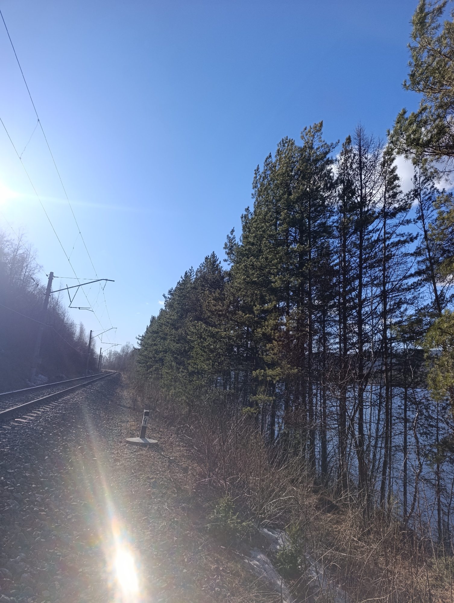The image depicts a section of railway track running alongside a body of water, likely a river or lake, under a clear, bright sky. The railway track is on the left, with associated overhead electrical infrastructure. To the right of the tracks is a line of trees, and beyond the trees, the water can be seen. The foreground vegetation appears to be mostly bare. The scene is flooded with sunlight. The specific location in Krasnoyarsk, Russia cannot be confirmed.