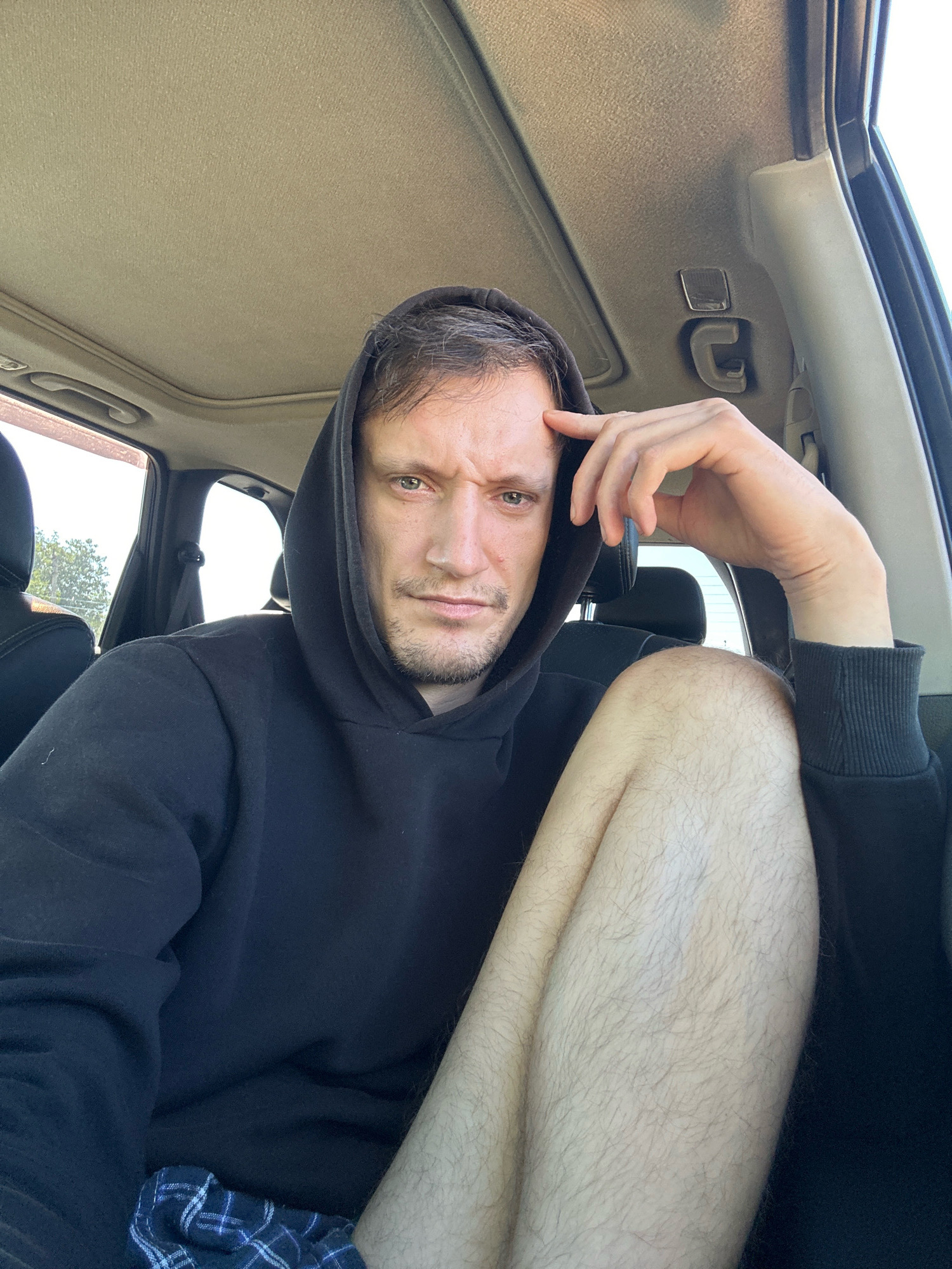 A man with grey hair and stubble, wearing a black hoodie, sits in the driver's seat of a car. His right hand is resting on his forehead, and his legs are crossed. The interior of the car, including the headliner and a portion of the dashboard, is visible. The background shows a bright, sunlit exterior with trees and a bright sky, suggesting an outdoor setting in Makiv, Ukraine.