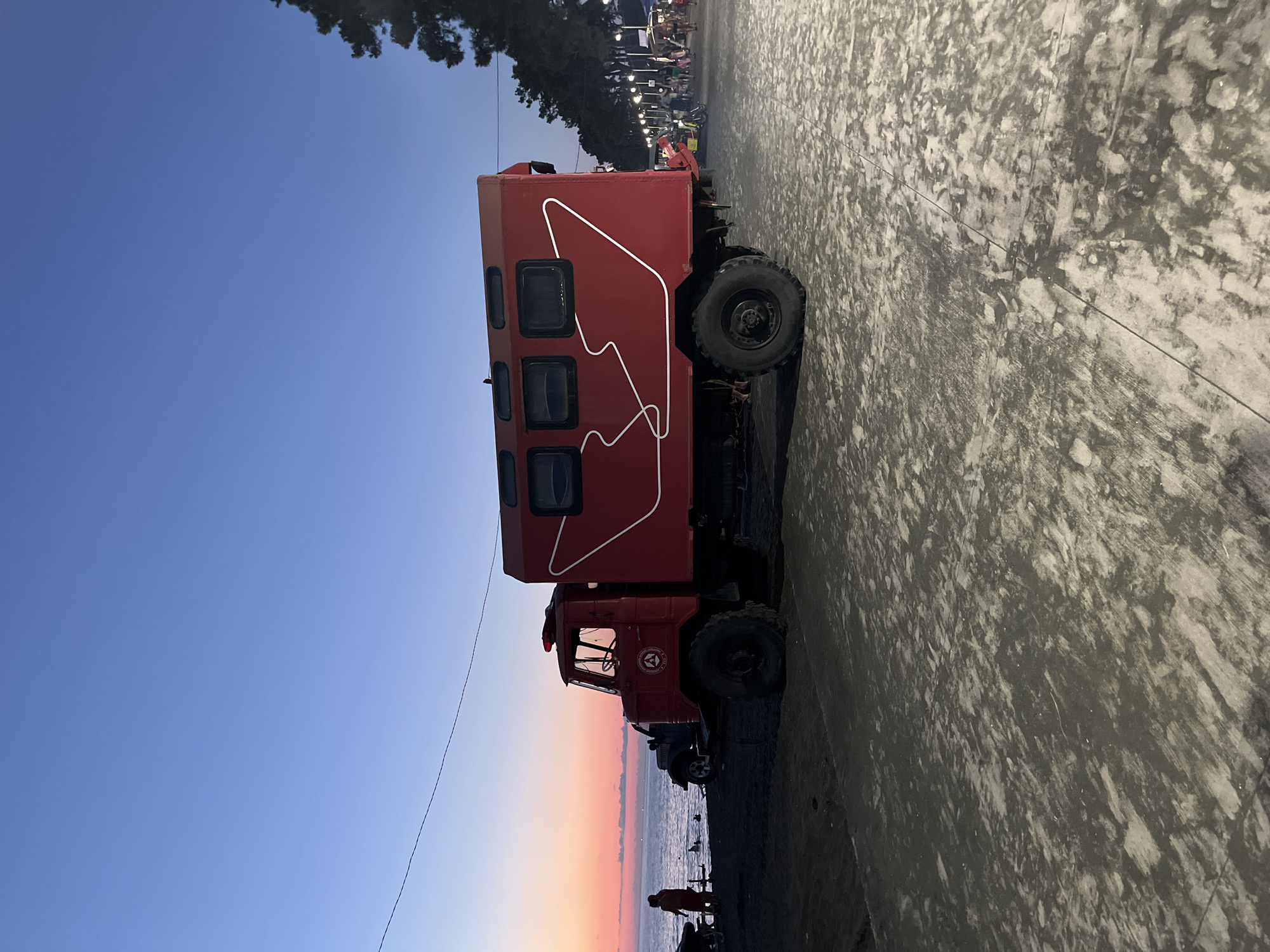 An outdoor, wide-angle, medium shot captures a bright red, utilitarian truck with a camper-style body parked on a dark, sandy surface. The truck is the dominant subject, positioned centrally and angled slightly towards the viewer's left. Its large, rugged tires suggest it is designed for off-road use. A white, abstract graphic, resembling a stylized line drawing, adorns the side of the camper body. The truck's cabin features a red beacon light on its roof.
Behind the truck, a body of water, likely the Black Sea, is visible under a sky transitioning from a soft pink and orange hue at the horizon to a deep blue overhead. The sun appears to have recently set or is about to rise. A few figures are present near the water's edge, including one person wearing an orange uniform. To the right of the truck, a paved walkway is illuminated by streetlights, with several people walking along it, some appearing to be children. Trees line the far side of the walkway.
The foreground is dominated by the dark, sandy ground, with patches of lighter colored material, possibly sand mixed with dirt or debris. A portion of the paved walkway is also visible in the lower right quadrant of the image. The overall scene suggests a coastal area, likely a promenade or beach area in Ureki, Georgia, during dusk or dawn.