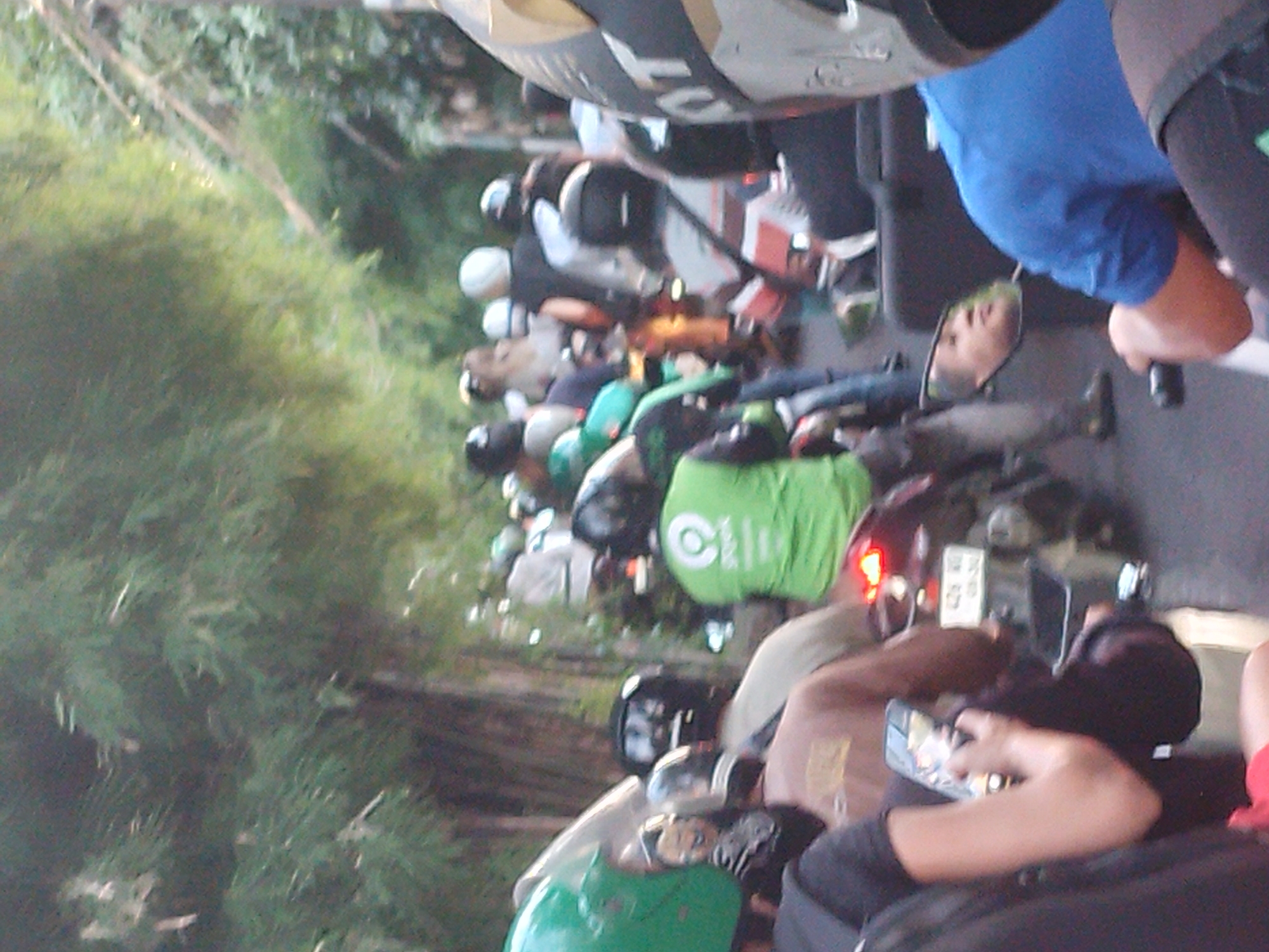 The image captures a bustling street scene in North Kuta, Indonesia, showing a dense queue or slow-moving traffic primarily composed of motorbikes. On the left side of the frame, lush green foliage suggests a natural or treelined environment bordering the road.
Numerous people are visible, each riding a motorbike and wearing a helmet of various colors including black, white, and brown. Many riders appear to be engaged in commuting or working, with several carrying bags or wearing jackets indicative of ride-hailing or delivery services. A prominent bright green bag with a white circular logo, strongly resembling the Gojek branding popular in Indonesia, is visible on one of the motorbikes. One person's face is clearly reflected in their motorbike's rearview mirror. In the immediate foreground, a rider is holding and looking at a smartphone.
The activity taking place is a typical urban traffic situation, with motorbikes lined up, possibly waiting at a traffic light or navigating a congested road. The lighting indicates it is daytime, with relatively soft light suggesting either morning or late afternoon rather than harsh midday sun. The scene reflects the common mode of transport and daily life in this region. Visible text includes the distinctive white circular logo on the green bag and some faint, partially obscured letters, possibly "OT" or "LOT," on a helmet or part of a motorbike near the top of the frame. An illegible license plate or screen is also visible on a motorbike in the mid-ground.