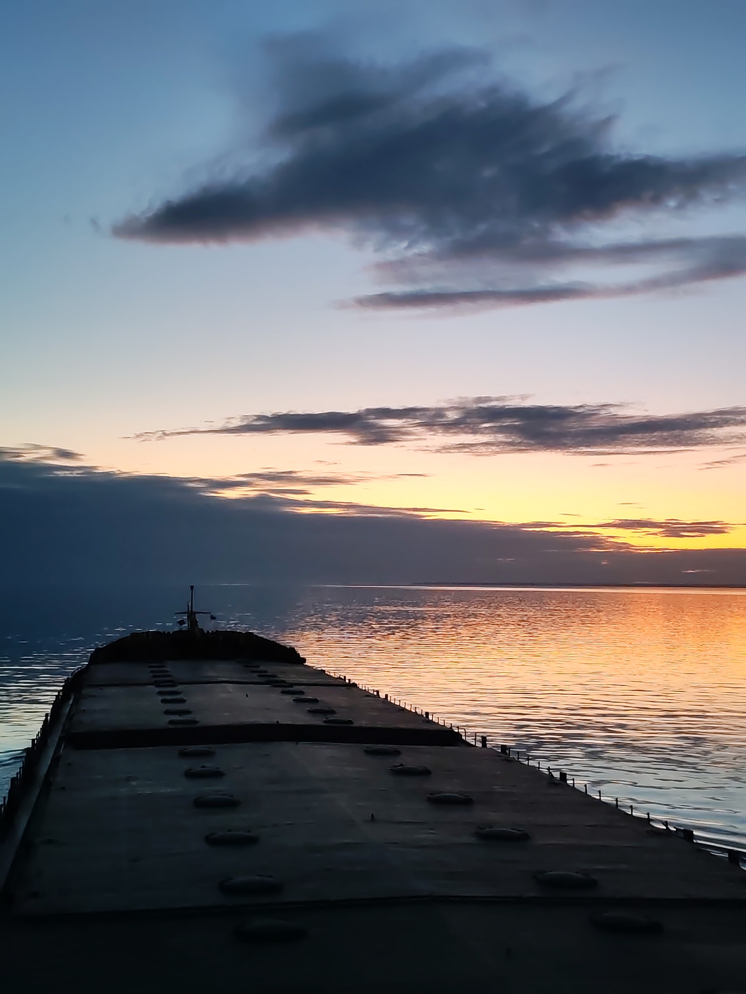 The image presents a serene twilight scene from the stern of a large, flat-decked vessel, likely a cargo ship or barge, on a vast body of water. The sky, a gradient of soft blue at the top, transitions into a vibrant display of orange and yellow hues at the horizon, indicating either a sunrise or sunset. Dark, elongated clouds stretch across the sky, creating a visual contrast with the brighter portions. This colorful sky is reflected on the calm water surface, which shimmers with the golden light, forming a path towards the distant horizon.
The vessel itself is primarily in silhouette, emphasizing its robust, industrial form. Its broad deck features multiple rows of evenly spaced, raised, oval or circular structures, which appear to be hatches or covers for cargo holds. Towards the far end of the visible deck, a small, dark structure with a mast or antenna is discernible. The water immediately surrounding the vessel shows gentle ripples. No people are visible in the frame. The overall atmosphere is tranquil and expansive. There is no discernible text present in the image. The location is contextualized as Unknown, Russia.