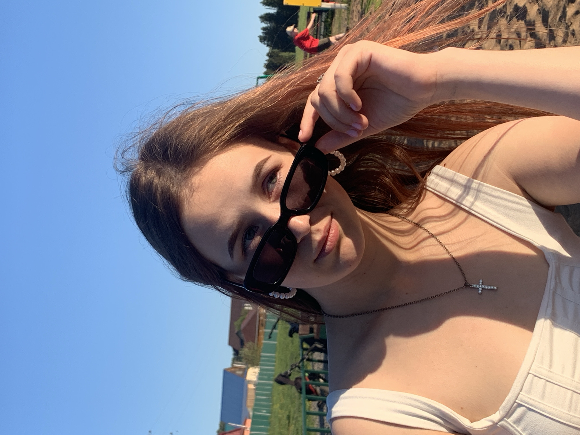 A young woman is photographed from a low angle against a bright blue sky. She is wearing a white top and dark sunglasses with pearl embellishments, and a delicate necklace with a cross pendant. Her hair is brown at the roots, transitioning to pink at the ends. She is holding the sunglasses on her face with her left hand, wearing a ring.
In the blurry background, there's greenery and some buildings, indicating an outdoor location, possibly a park or recreational area. In the distance, a figure in a red shirt appears to be suspended in the air or mid-motion, possibly engaged in some form of activity, like gymnastics, or playing. Behind that figure are trees. The overall scene suggests a sunny day in a public park. It is not possible to verify the location as the city of Фоки, Russia.