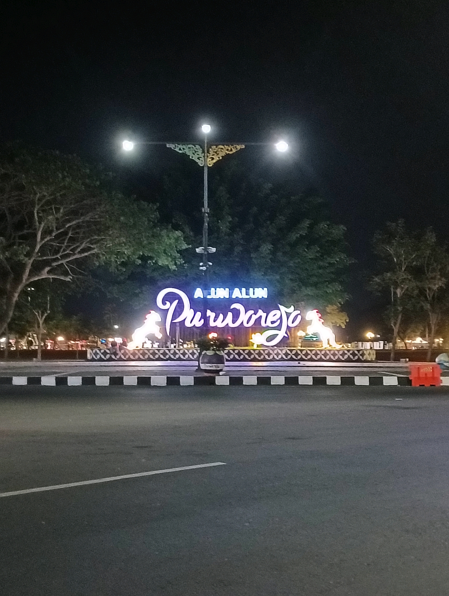 The image shows a night scene in the city of Purworejo, Indonesia, featuring the "Alun-Alun Purworejo" landmark. The central focus is a large, illuminated sign that spells out "ALUN ALUN Purworejo" in stylized, glowing letters. Flanking the sign are two illuminated figures, appearing to be lions or mythical creatures, made of white light. The sign and figures are erected on a patterned base. In the foreground, there is a paved road with a black and white checkered curb. Behind the landmark, trees are visible, their leaves illuminated by ambient light and the glow from the sign. A street lamp with decorative metalwork is positioned above the sign. The overall atmosphere is one of nighttime illumination in a public space. There are no people visible in the image.