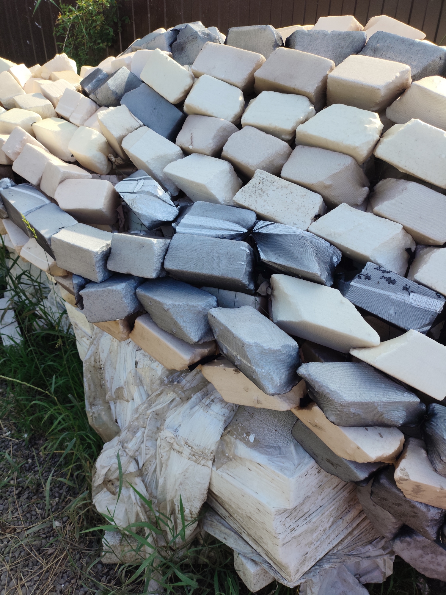The scene depicts a large, irregularly stacked pile of diverse blocks situated outdoors in Prigorodnyi, Russia. The pile consists predominantly of two types of materials: numerous light-colored, off-white to cream-colored blocks with textured, porous, or slightly damaged surfaces, consistent with foamed insulation or lightweight construction material; and various grey-colored blocks that appear to be wrapped in a dark, fine-mesh netting. Some of these netted blocks exhibit tears or exposed underlying material. Interspersed are a few yellowish or beige blocks.
The blocks are arranged haphazardly, forming a substantial mound that extends from the foreground towards a dark brown, possibly wooden plank fence in the background. Green foliage, suggestive of bushes or small trees, is visible behind the fence. The ground surrounding the base of the pile consists of dark earth, scattered gravel, and patches of green grass. Crumpled white plastic sheeting is visible at the very bottom and around the base of the stack. No individuals, animals, or explicit actions are depicted. The lighting indicates an outdoor daytime setting with clear sunlight creating shadows on the blocks.