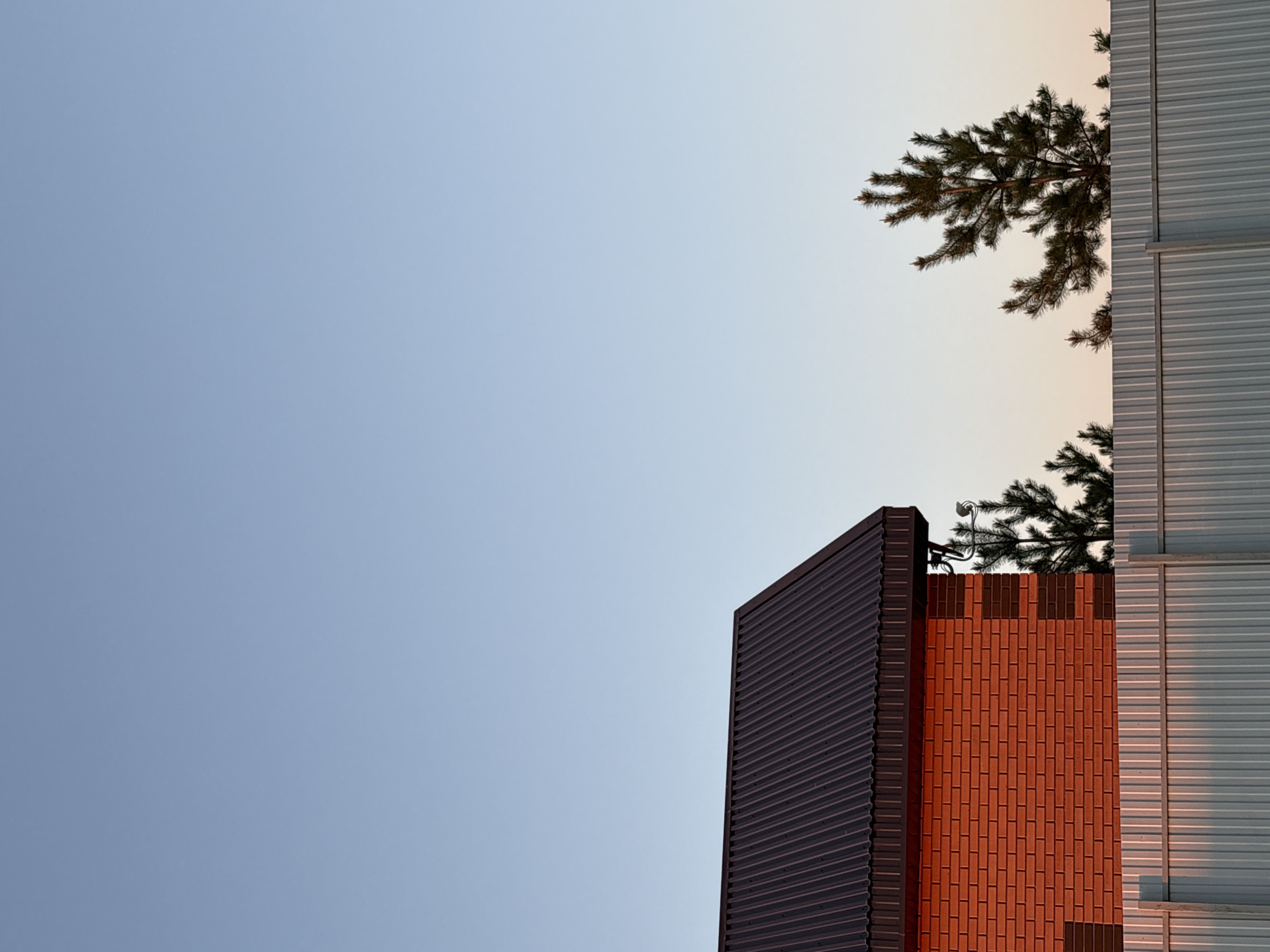 A low-angle shot of buildings in Кырныш, Russia, reveals a blue sky occupying the upper left portion of the frame, transitioning to a soft orange hue in the upper right. In the lower right, a building with corrugated metal siding stands next to a brick structure, both partially obscured by a pine tree branch. A dark, corrugated building is on the left. The scene is still and devoid of any visible human activity.