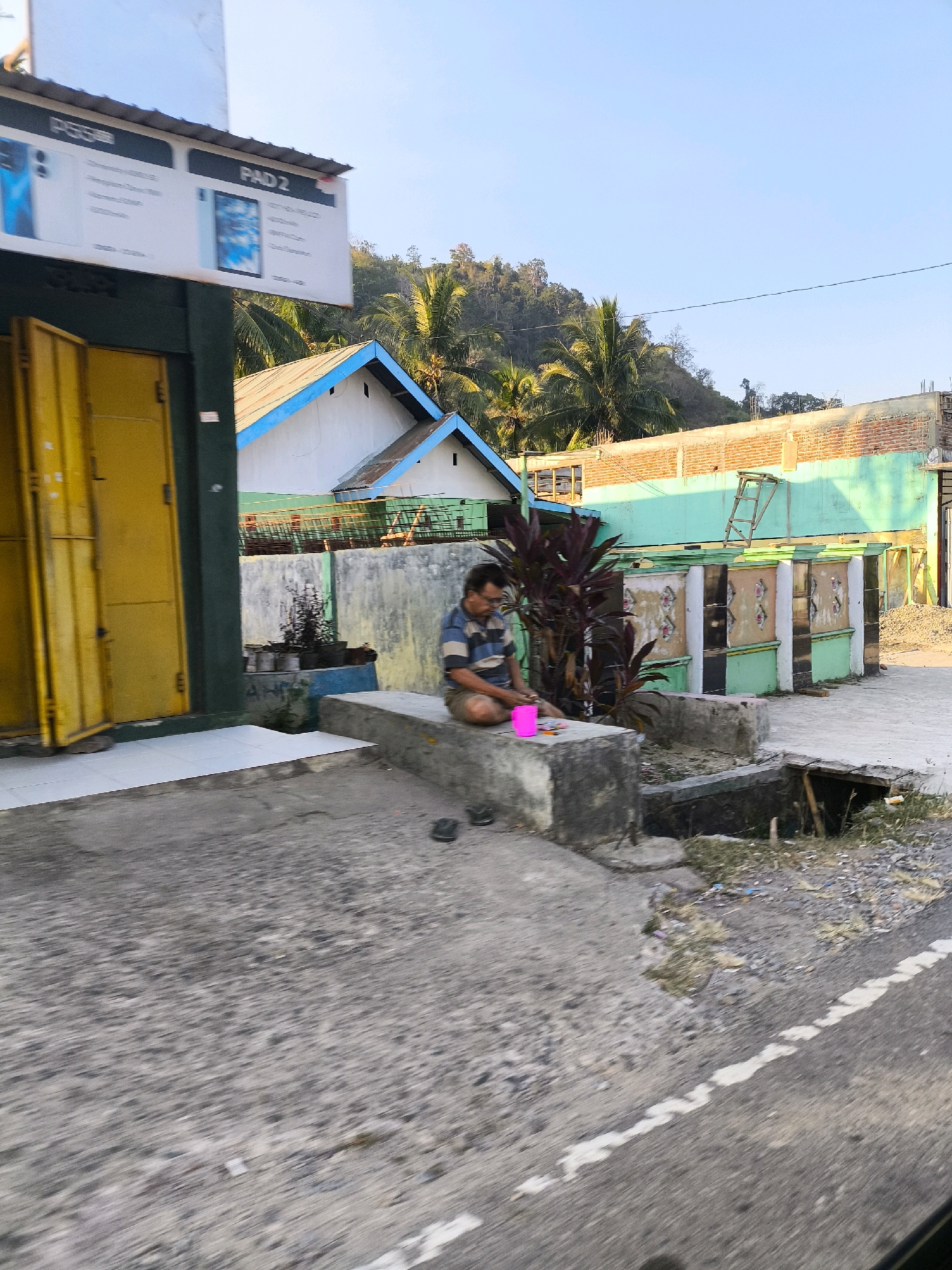 The image shows a street scene in Dulupi, Indonesia. In the foreground, a man in a striped shirt and shorts sits on a concrete ledge, looking down at a bright pink object on the ledge in front of him. He is seated near a building with a yellow door and a sign that reads "PAD 2". In the background, there are several buildings with colorful facades, palm trees, and a hillside covered in foliage. The sky is a clear blue, suggesting it is daytime. The overall impression is a snapshot of everyday life in a tropical urban environment.