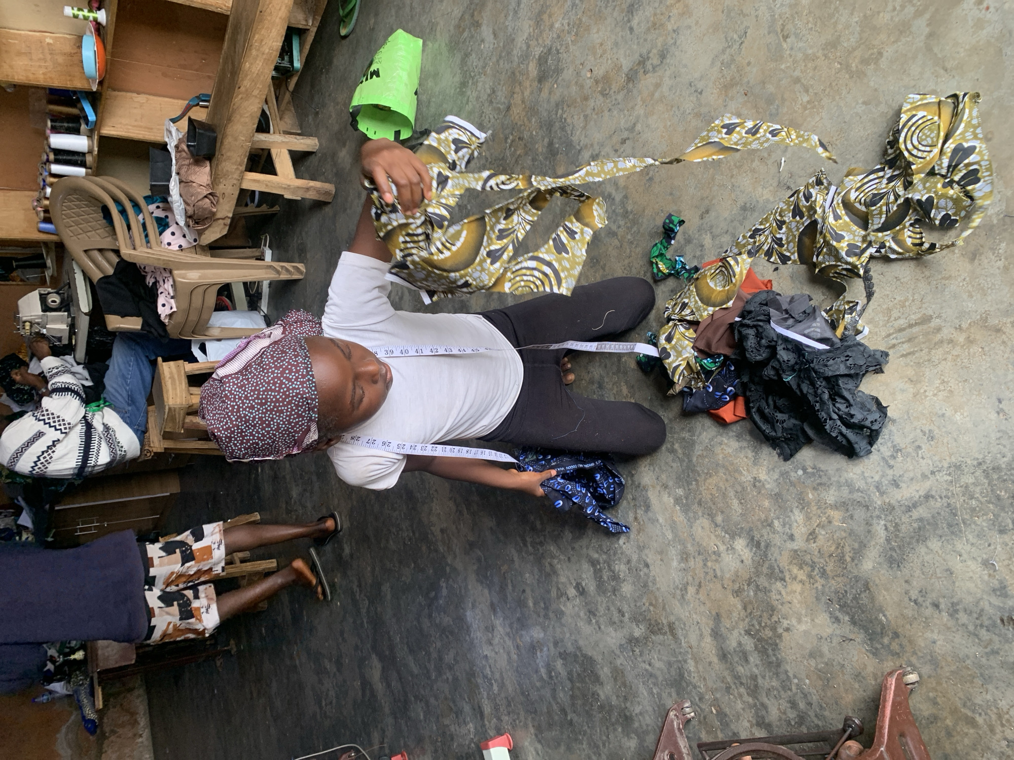 An overhead shot captures a person, likely a woman, sitting on a concrete floor in what appears to be a tailoring workshop or home-based business in Jos, Nigeria. She is wearing a white t-shirt, black trousers, and a patterned head covering. A measuring tape, displaying numbers from '12' to '28', is draped around her neck, indicating her role in dressmaking or tailoring. She holds a piece of olive green, black, and white patterned fabric, seemingly in the process of working with it.
Scattered on the floor around her are various fabric scraps, including more of the patterned material, a blue patterned fabric, black lace, and other colored pieces. To her left, the lower half of another person is visible, standing on the concrete floor, wearing patterned shorts and a dark top.
The setting includes rustic wooden structures, possibly shelves or workbenches, against a wall. Spools of colorful thread are neatly arranged on these shelves. A light brown plastic chair and parts of what appears to be a sewing machine are also visible. A bright green plastic bag, with the letter "M" visible on its side, rests near the main subject. The floor is a worn, textured concrete surface. The scene conveys an industrious atmosphere, focusing on the craft of textile work.