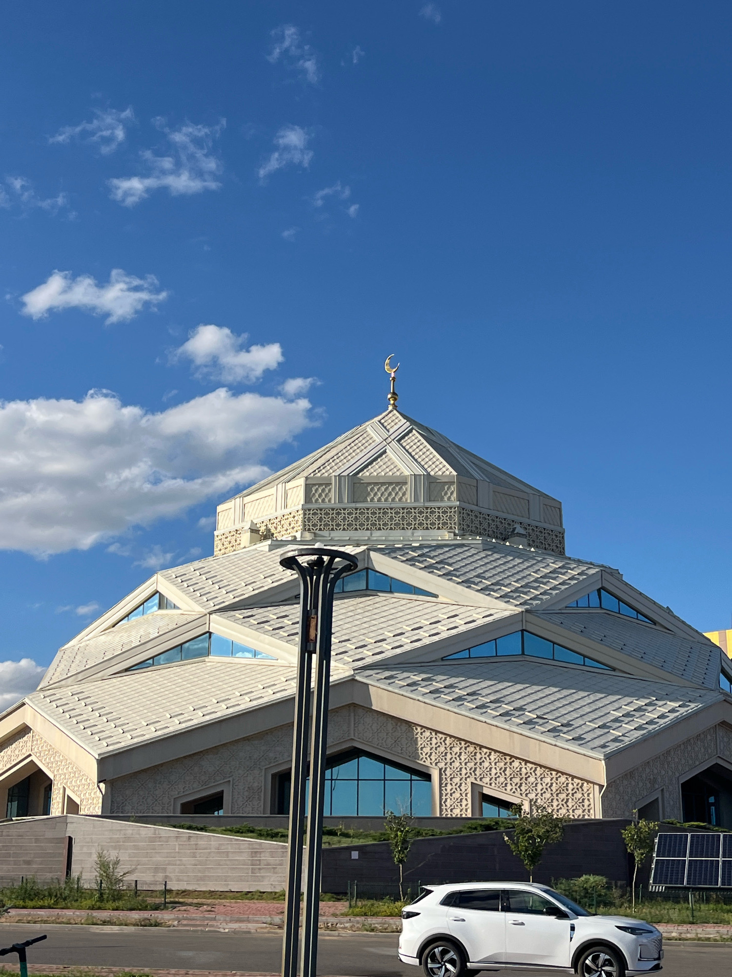 The image captures a white car parked on a street in front of a modern mosque in Astana, Kazakhstan. The mosque features a distinctive geometric design with a prominent dome topped with a golden crescent moon. The building is constructed with light-colored stone and features angular rooflines with triangular blue-tinted windows. In the foreground, a lamppost stands on the side of the road, and a portion of a scooter is visible on the left. The background shows a bright blue sky with scattered white clouds. Solar panels are visible to the right of the mosque. The overall scene depicts a contemporary urban landscape in Astana under a clear sky.