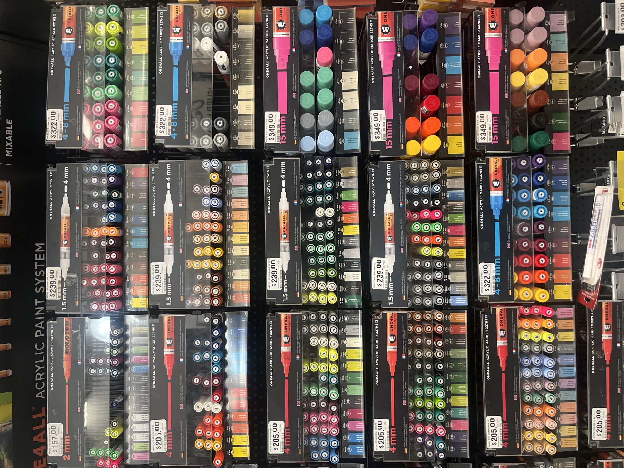 The image shows a display of ONE4ALL acrylic markers in various sizes and colors, neatly arranged on shelves in a retail setting. The markers are priced between $157.00 and $349.00, depending on the size and type. The environment appears to be a well-lit aisle of an art supply store or a similar retail establishment. The brand names "ONE4ALL" and "MOLOTOW" are visible. There are no people visible in the image. The markers are the primary subjects, and the prices are displayed on the packaging. No specific location in Puebla, Mexico, is identifiable from the visual information provided.