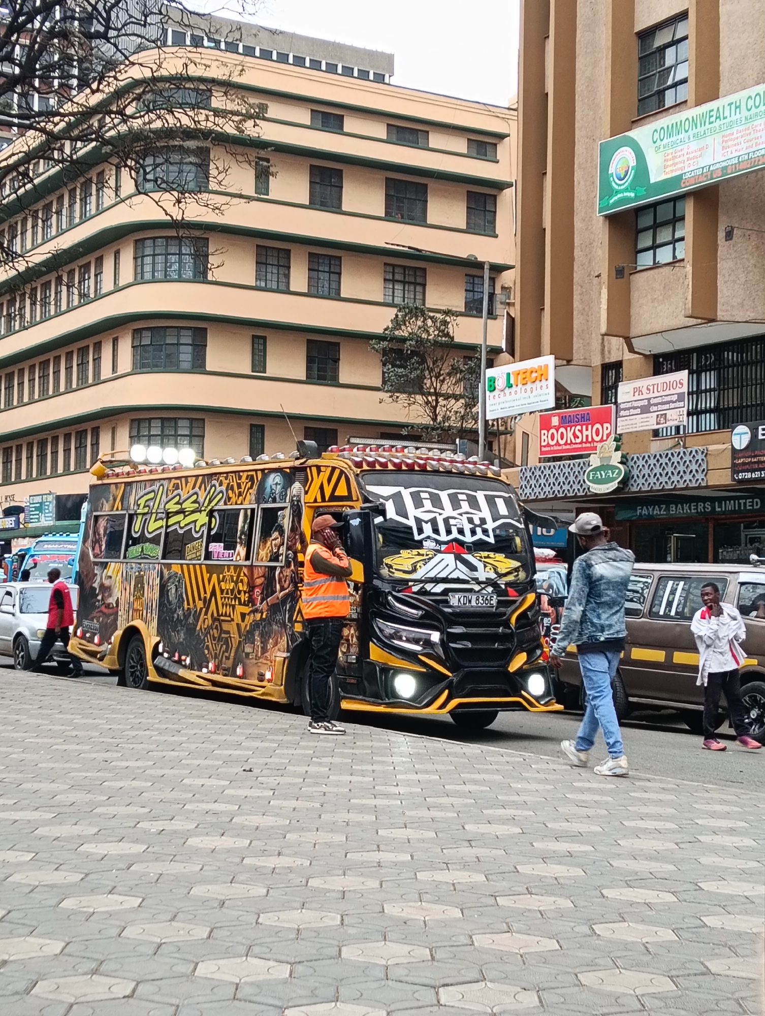This is a street-level, medium shot photograph taken in downtown Nairobi, Kenya, likely on a street with commercial buildings. The focal point is a vibrantly decorated matatu, a public transport minibus, parked on the street. The matatu is predominantly yellow and black, with extensive graffiti art and various advertisements displayed on its sides. It has numerous lights on its roof and at the front. In front of the matatu, a man wearing an orange safety vest and a cap is standing, appearing to be a conductor or attendant. To the right of the matatu, another man in a denim jacket and jeans walks away from the camera. Further to the right, a matatu is partially visible. In the background, there are multi-story buildings, including a light peach-colored building with rounded corners and green trim, and a tan building with a prominent sign for "COMMONWEALTH COLLEGE OF MEDICAL & RELATED STUDIES." Other signs for businesses like "MAISHA BOOKSHOP" and "FAYAZ BAKERS LIMITED" are also visible. The ground is paved with hexagonal interlocking stones. The overall atmosphere suggests a busy urban environment.