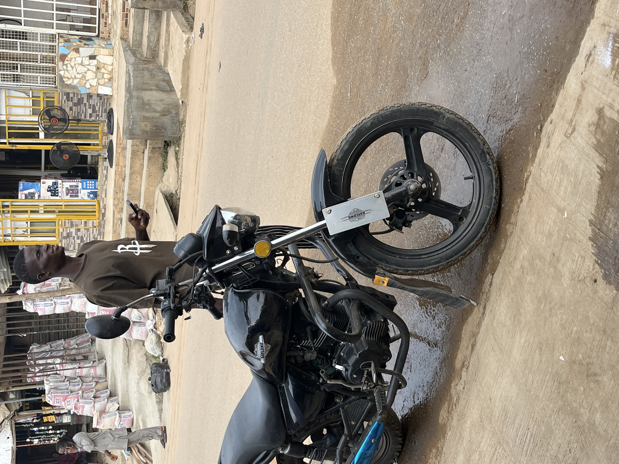 The image displays an outdoor commercial scene in what is noted as Abuja, Nigeria. A black motorcycle is parked on a concrete surface, with a young man standing beside it.
The motorcycle, identified by the "DAYLONG" brand on its front mudguard and fuel tank, is predominantly black with chrome accents. It features a single front disc brake and a black spoked wheel. The man, wearing a dark olive-green t-shirt with a white stylized logo (resembling "BS"), holds a black smartphone in his right hand and looks off to the left with a neutral expression.
In the background, substantial stacks of cement bags, some marked "3X", are visible, indicating a building material supply point. Adjacent to this, a yellow metal railing fronts an area where two black standing fans are positioned. Behind these, a display of electronics, possibly speakers and radios, can be seen on shelves. Further back, concrete steps lead up to a building partially adorned with small mosaic-like tiles. Another person in light traditional attire stands near the cement bags, looking downwards, while the lower body of a third individual is partially visible further left.
The scene is well-lit, suggesting daytime. The ground consists of concrete and dirt, with some damp areas. The overall impression is that of a typical bustling street or market environment in an urban or semi-urban setting.
