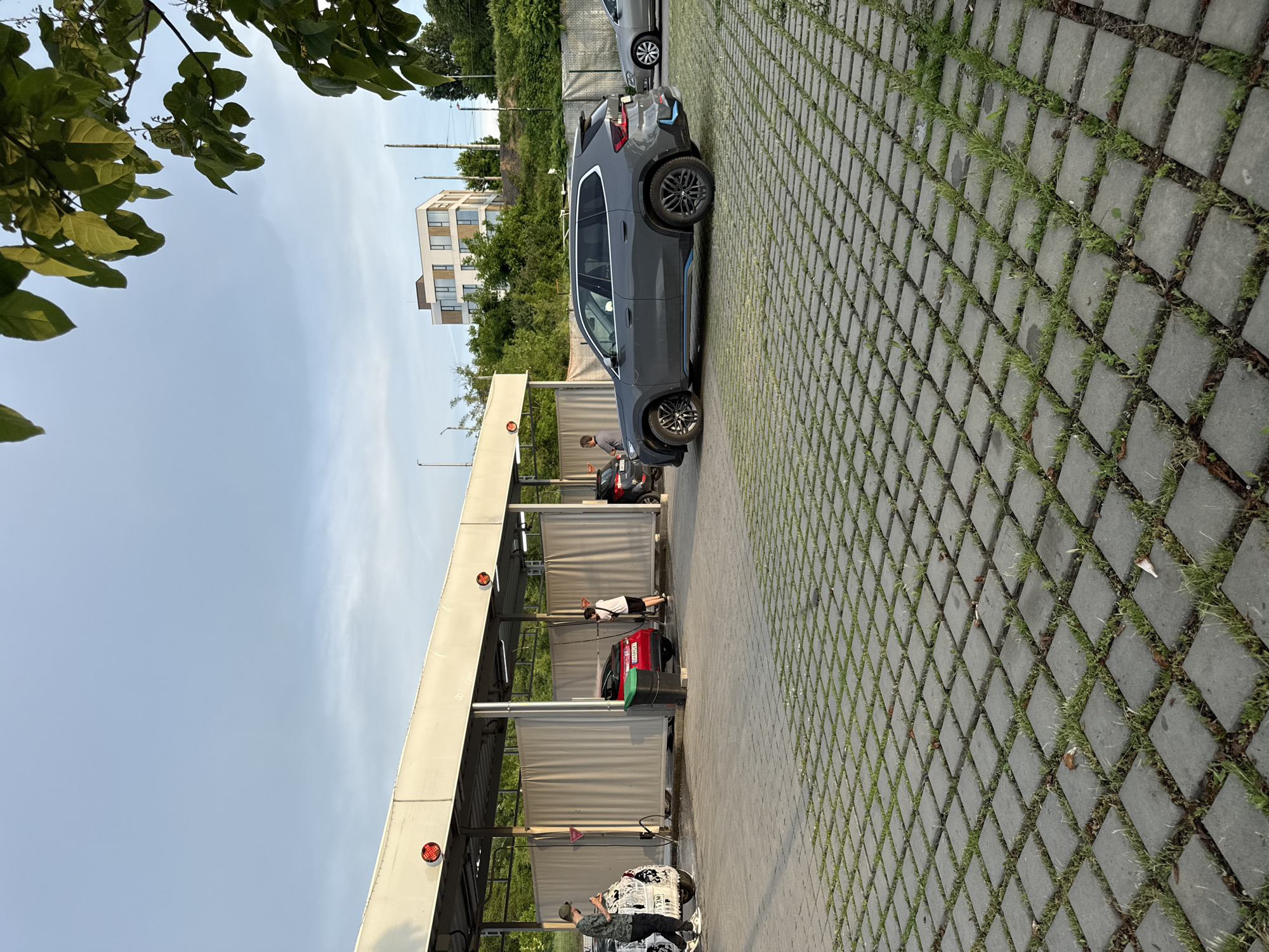 The image depicts a self-service car wash in Pleven, Bulgaria. Several people are present, seemingly washing or detailing vehicles. The facility features multiple bays under a white roof, each equipped with curtains. Two cars are within the bays, and a gray SUV is parked nearby. The ground is paved with square concrete blocks with grass growing between them. In the background are trees and a modern building. The sky is overcast with light clouds. A tree is in the upper left.