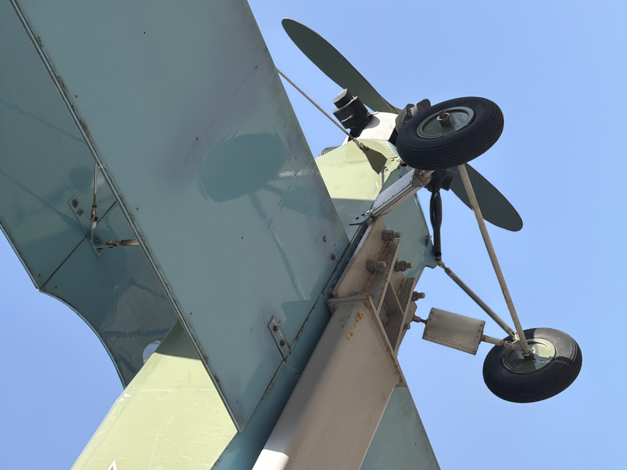 The image displays the underside of a vintage Polikarpov Po-2 biplane monument, suspended against a clear, unobstructed blue sky. The aircraft's lower wing, painted in a faded light blue-green, occupies the upper and right portions of the frame, showing signs of weathering and minor rust along its seams and rivets. An olive-green structural section, possibly part of the fuselage or an upper wing support, is visible to the upper left. A heavy, light grey rectangular support beam, secured by multiple large bolts, extends from the main structure. Below this, the landing gear is prominent, featuring two black rubber tires with olive green wheel hubs, connected by metal struts. A two-bladed propeller, also olive green, is partially visible behind the rightmost wheel, adjacent to what appears to be part of the engine block. Strong sunlight casts defined shadows across the aircraft's surfaces. No individuals or other environmental elements are present. This static display is located in Krasnoye Selo, within the city of Saint Petersburg, Russia.