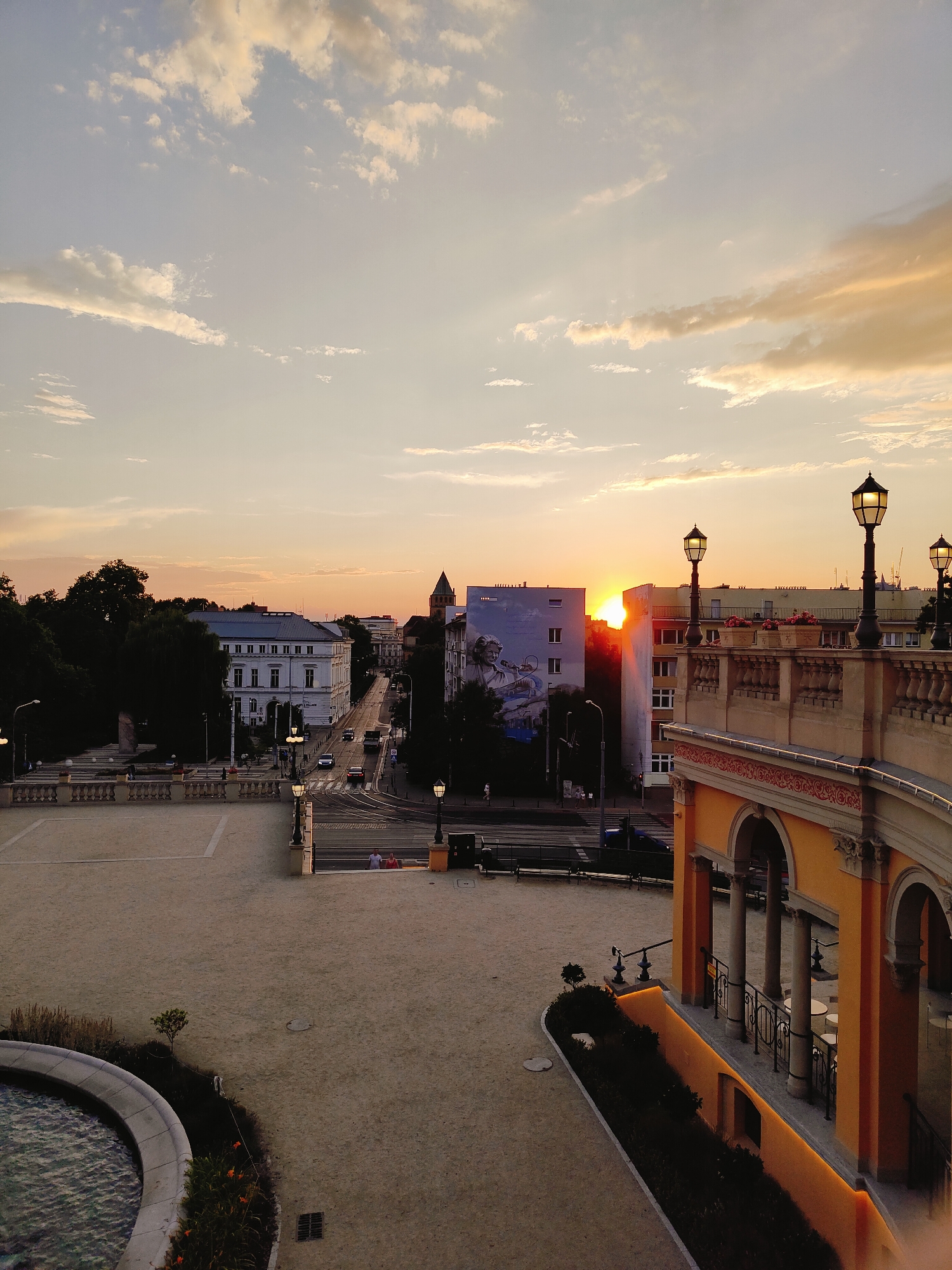The image captures a sunset over the city of Wrocław, Poland. The view is from a high vantage point, looking down at a city square and street with buildings and trees. The sky is filled with warm hues of orange and yellow, with scattered clouds illuminated by the setting sun.
In the foreground, a beige-colored paved area, likely a public square, is visible. To the right, a section of an ornate building with arches and decorative railings, painted in a vibrant orange and yellow, stands out. This structure appears to be a balcony or an elevated walkway. Lampposts with old-fashioned designs are placed along the edge of this elevated area.
Further into the scene, a street lined with buildings runs into the distance. One prominent building is white with many windows, and across the street, a building features a large mural of a woman. Vehicles are seen on the street, indicating it is an active thoroughfare. Trees are scattered throughout the cityscape, adding greenery to the urban environment.
In the lower left corner, a portion of a circular fountain or pool is visible, with water in it. The ground is covered with sand or gravel in some areas, and paving stones in others. The overall atmosphere is serene, with the warm light of the sunset casting a golden glow over the scene. There are a few people visible in the distance, walking or standing. The scene appears to be a public space within the city, possibly near a cultural or historical landmark.