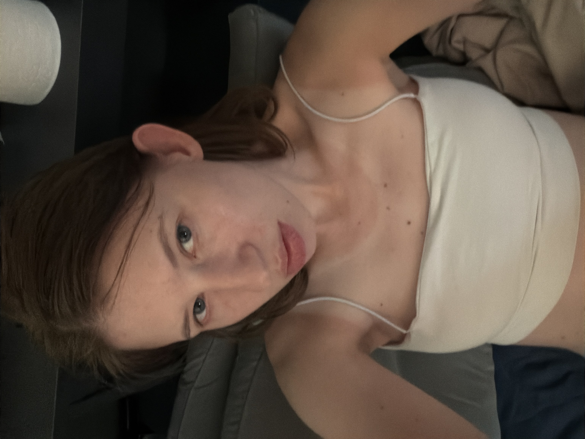 A young woman with brown hair is lying down, looking at the camera. She is wearing a white tank top. The background is dark and out of focus, but appears to be indoors, possibly a bedroom. There is a roll of toilet paper visible in the upper right corner of the image.