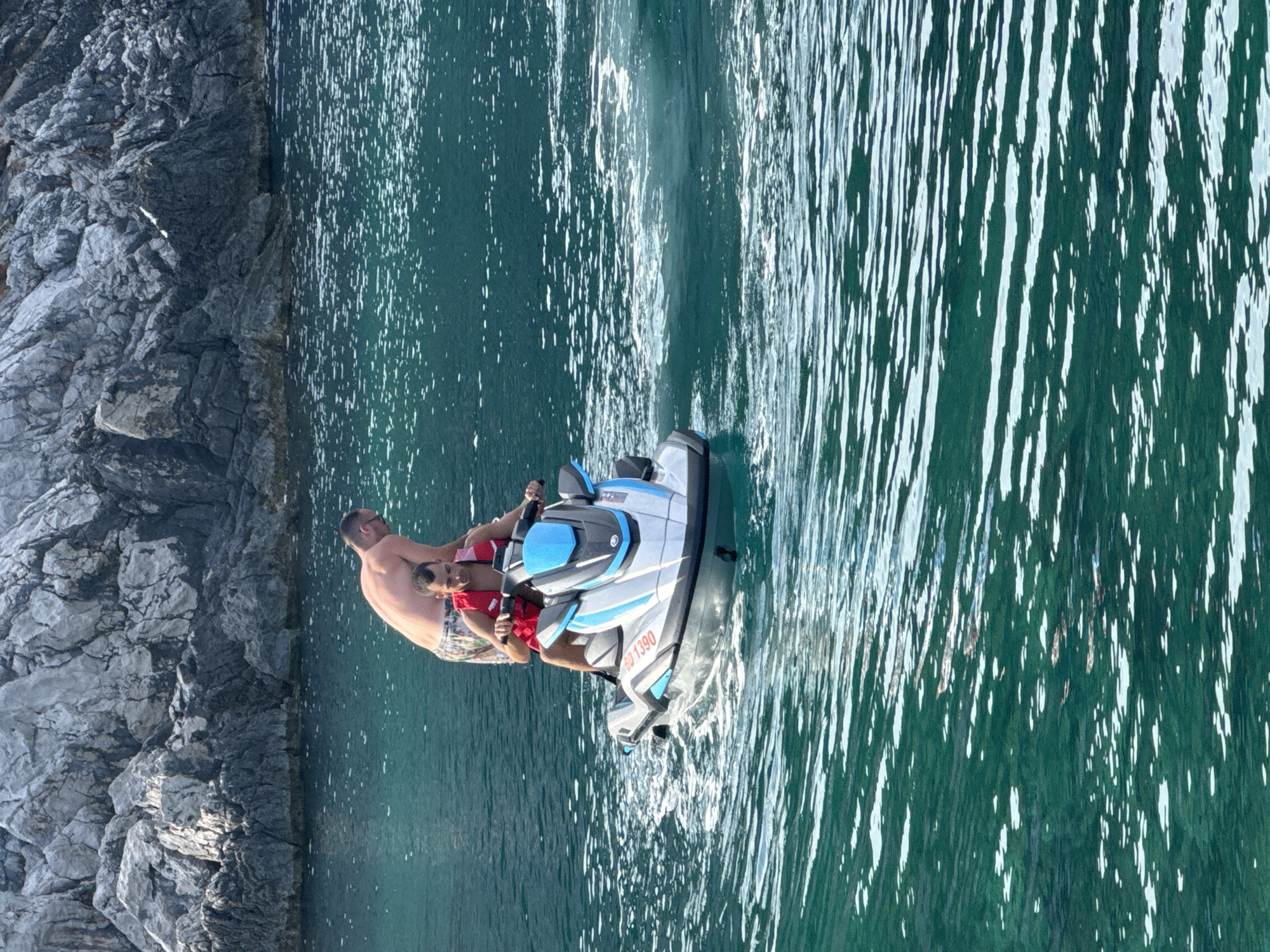 Two men are on a jet ski in the emerald green water off the coast of Dubraljevina, Montenegro. The man in front is driving the jet ski and wearing a red life vest. The man behind him is shirtless and wearing patterned shorts. In the background, a rocky cliff face rises from the water. The water is choppy, with white spray trailing behind the jet ski.