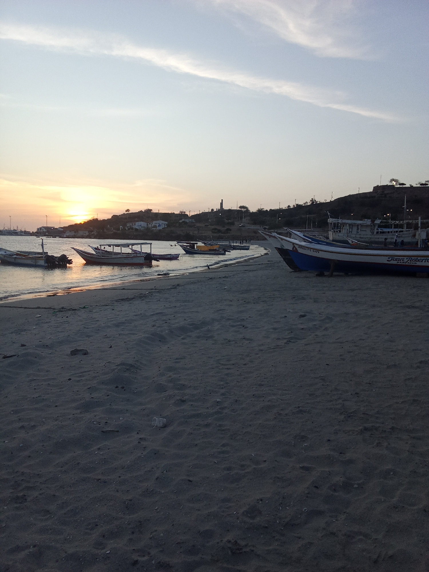 The image depicts a beach scene in Juan Griego, Venezuela, at sunset. Several small fishing boats are beached on the sand, with others anchored near the shore in the calm water. The sun is setting on the horizon, casting a warm glow on the sky and water. In the background, there are buildings and vegetation on a hill. The foreground consists of sandy beach with some small rocks and debris visible. There are no people discernible in the image.