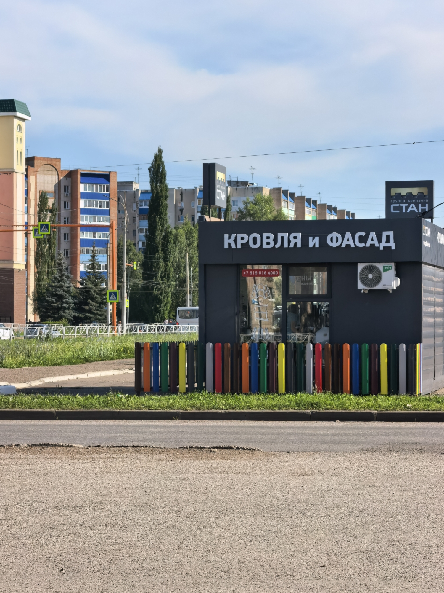 In Sterlitamak, Russia, a low-rise commercial structure with the sign "КРОВЛЯ И ФАСАД" (Roof and Facade) is prominently displayed. The structure is dark gray and features a glass storefront with reflections of buildings. An air conditioning unit is mounted on the exterior wall. In front of the storefront, a low fence made of brightly colored vertical poles in shades of red, orange, yellow, green, blue, and gray is visible. The structure is situated adjacent to a grassy area and a paved road. In the background, a row of multi-story apartment buildings with blue and white accents lines the horizon, under a clear blue sky with scattered clouds. Traffic lights and pedestrian crossing signs are also visible in the mid-ground.
