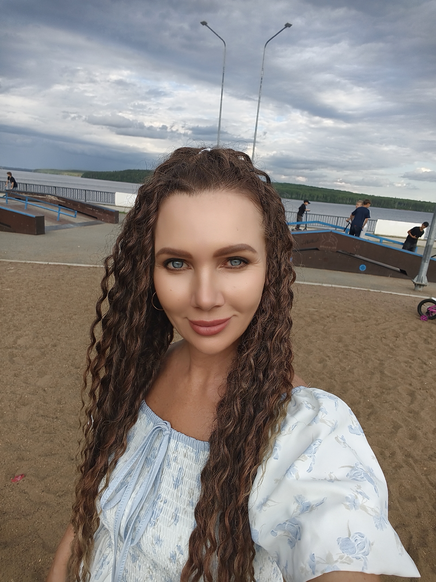 The image is a selfie taken by a woman with long, curly brown hair and blue eyes, wearing a white and blue floral print dress. She is standing on a sandy area in front of a body of water and a skatepark. In the background, there is a cloudy sky with a hint of blue, and several people are visible at the skatepark, some on scooters. The location is identified as the embankment of the Kama River in Chaikovsky, Russia. The scene appears to be a public recreational area, possibly a park or waterfront promenade. The woman is smiling at the camera, and the overall mood of the image is casual and outdoor-oriented.