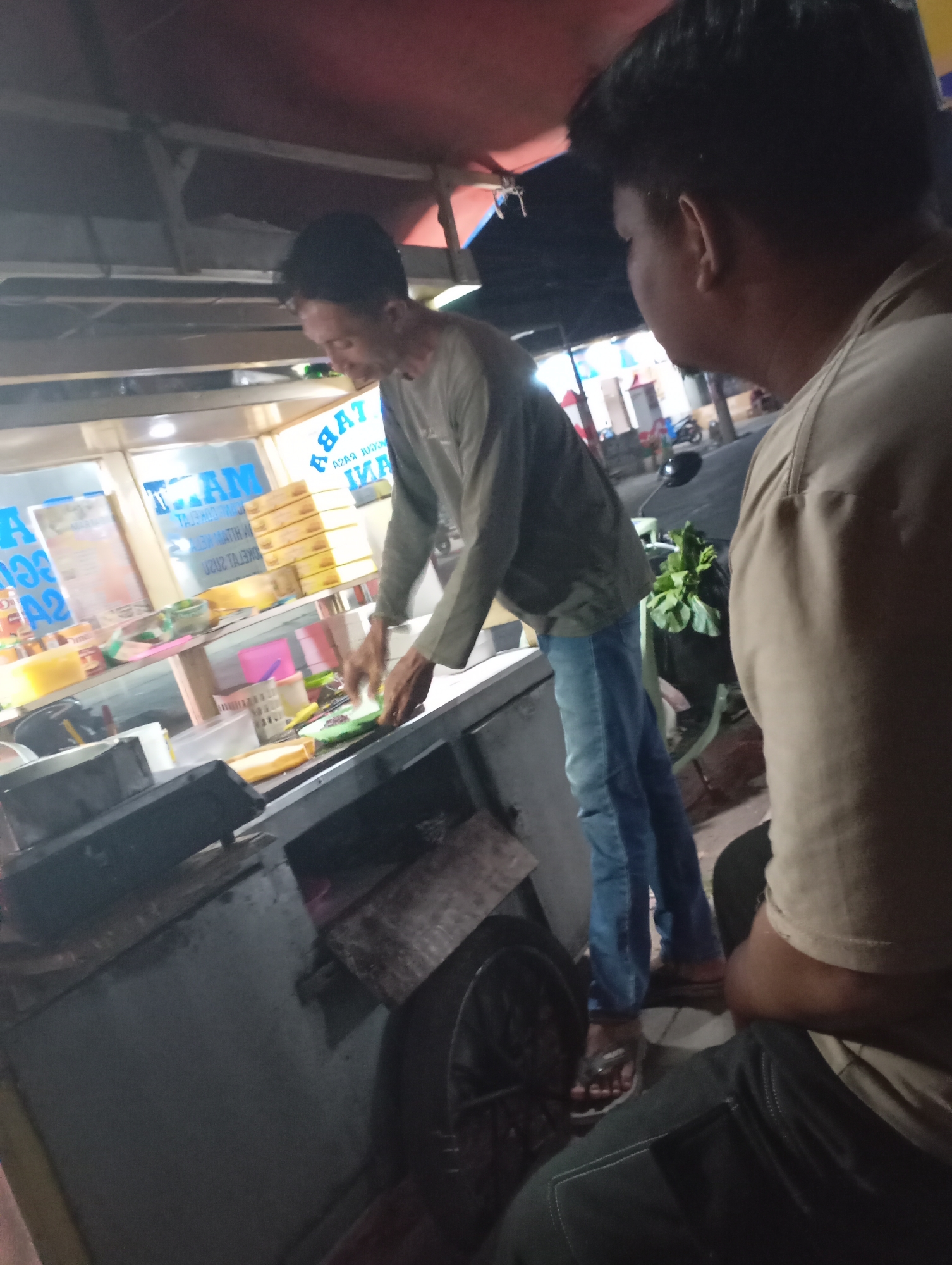 The image depicts an outdoor food stall at night in Kecamatan Haurgeulis, Indonesia. Two men are present. One man, with dark hair and wearing a grey long-sleeved shirt and blue jeans, is standing and actively preparing food, holding a green item with his hands over a counter. He is facing a display case. The second man, seen from the back and right side, has dark hair and wears a light brown or beige shirt. He is seated.
The food stall features a metal counter with various items on it, including containers, what appears to be food items, and possibly a cooking apparatus. Behind the counter, there are shelves with packaged goods, some of which have text that appears to be in Indonesian. A bicycle is visible in the background, with a bunch of green vegetables on its seat. The environment is dimly lit, suggesting it is nighttime, and the ground appears to be paved. The overall scene suggests a typical roadside food vendor operation.