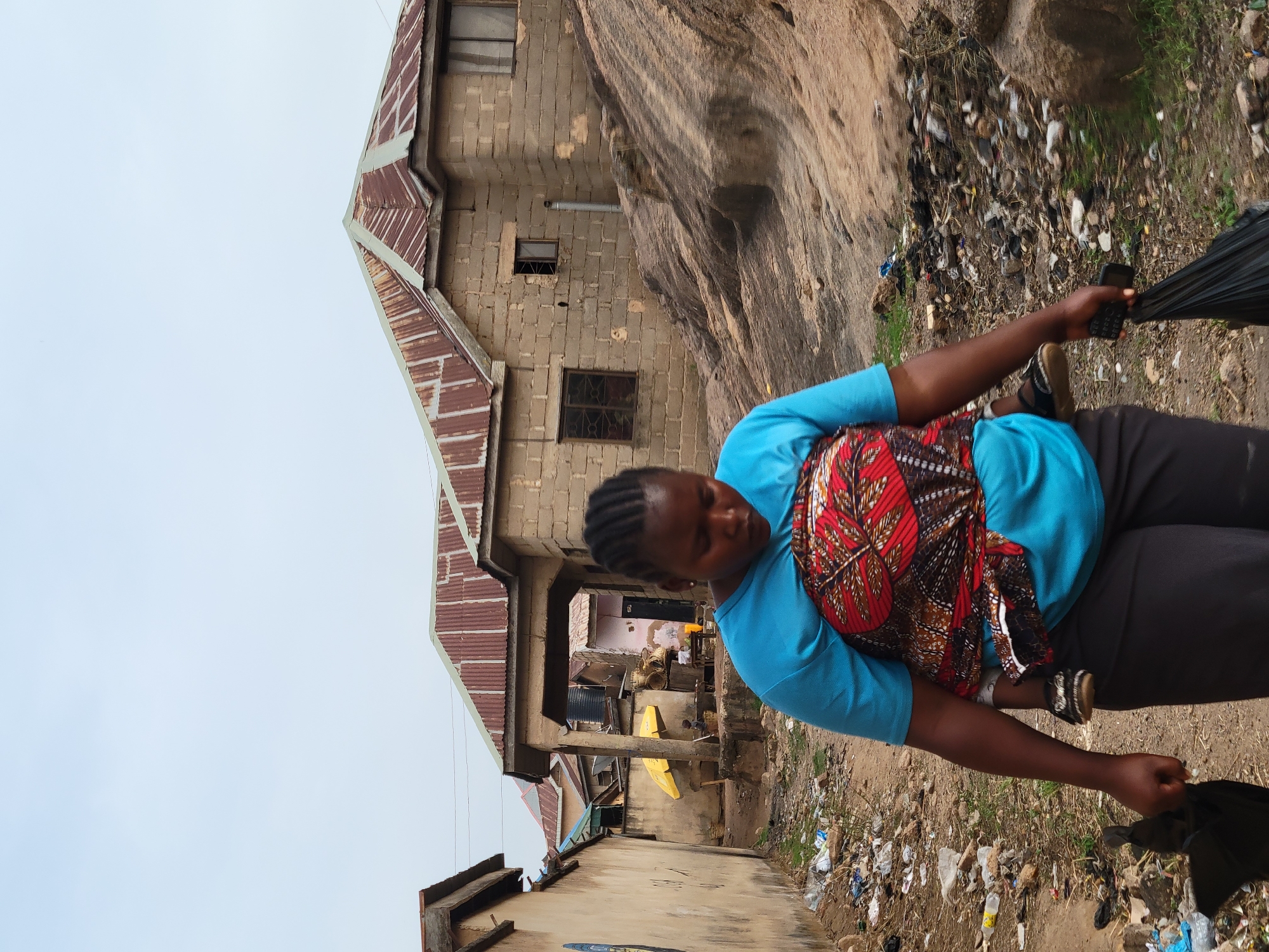 A medium shot depicts a woman with a child on her back, likely in Jos, Nigeria. The woman, wearing a light blue top and dark pants, is holding a black object in her right hand and a cell phone in her left. She has braided hair and is looking down. The child is secured to her back with a patterned cloth.
Behind her is a partially constructed brick building with a rusty metal roof. Adjacent to the building is a rock formation. The ground is littered with trash. The sky is overcast.