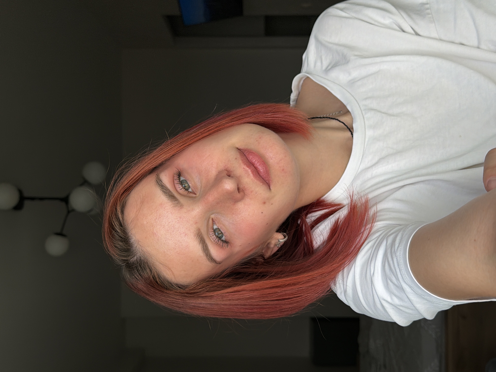 A woman with reddish-brown hair and fair skin is wearing a white t-shirt and gazing at the camera. She is indoors. A light fixture with multiple spherical bulbs is visible in the background, along with neutral-colored walls. The location is Naberezhnye Chelny, Russia.