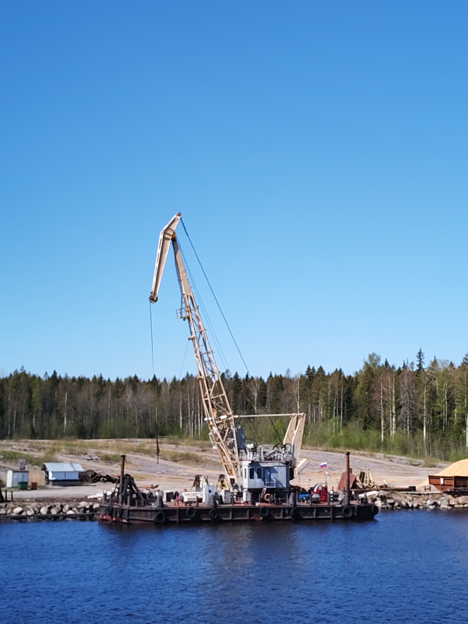 The image depicts a crane barge moored at a harbor in Davydovskaya, Russia. The barge is equipped with a lattice boom crane, and it is positioned near the shoreline. A forest is visible in the background. The sky is clear and blue. There are no people visible in the scene.