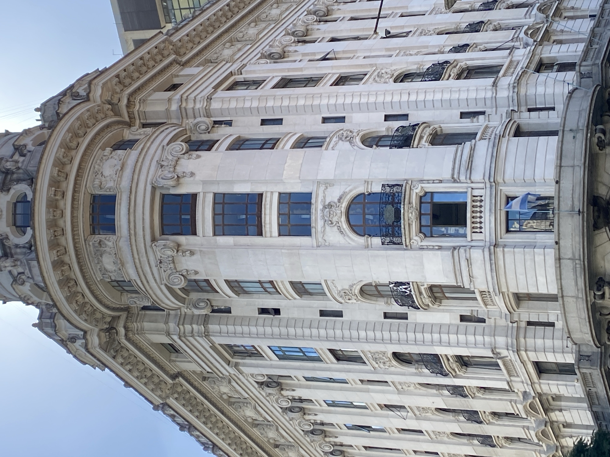 This image captures a close-up, angled view of a grand, ornate building in Buenos Aires, Argentina, under clear daylight. The primary subject is the building itself, characterized by its intricate, light-colored facade, likely stone or stucco, showcasing classical architectural influences.
The building features multiple levels with a rich array of decorative elements. Prominent details include a heavily embellished cornice line with detailed carvings, swags, and what appear to be sculptural figures or gargoyles. Below this, there are numerous windows of varying shapes: some are large rectangular panes, while others are gracefully arched. Many windows are recessed and framed by decorative moldings and pilasters. Several windows on different floors are adorned with elegant, dark-colored wrought iron balconies or balustrades with intricate scrollwork.
A notable feature is the curved or rounded section of the building, possibly a corner or a central bay, which adds to its distinctive silhouette. This section also boasts arched windows and decorative elements that mirror the rest of the facade. The detailed craftsmanship is evident in the friezes, cornices, corbels, and other sculptural embellishments that adorn almost every surface.
Through one of the windows, a blue and white striped flag, consistent with the flag of Argentina, is partially visible, providing a clear location cue. The sky above is bright and clear, indicating fair weather during the day. No people or specific activities are discernible, allowing the focus to remain entirely on the architectural splendor of the structure. No visible text is present on the building.