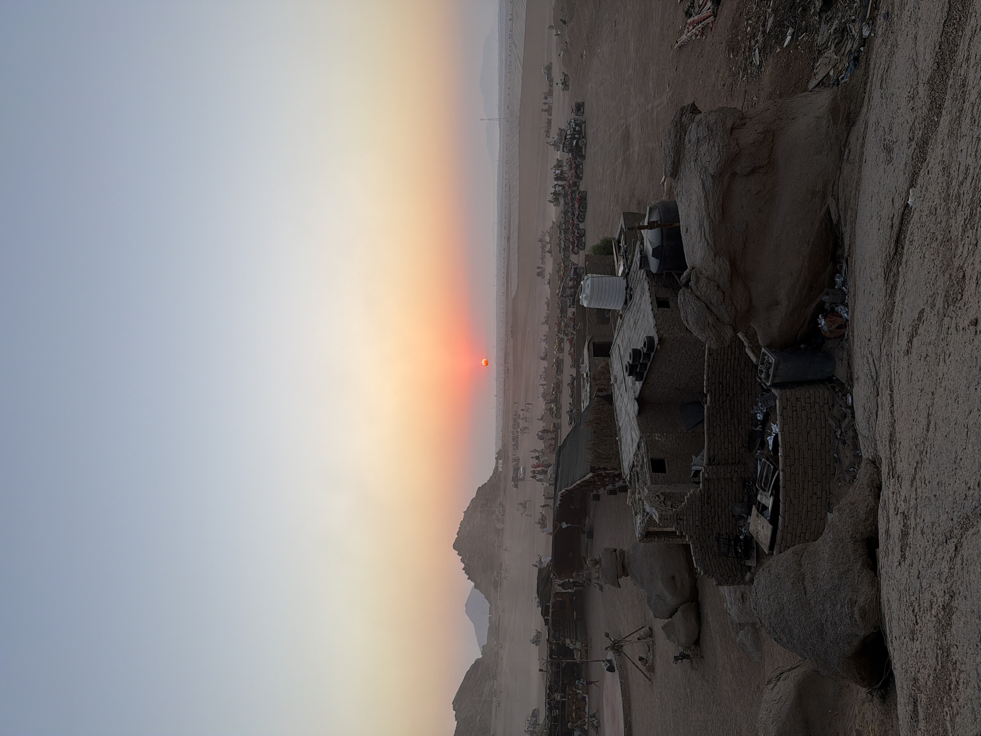 The image captures a desert landscape during sunset in Sharm El Sheikh, Egypt. The sky transitions from a pale blue at the top to warm hues of orange and pink near the horizon, where a bright orange sun is visible. In the foreground, rustic, low-rise structures made of brick and thatch are scattered across the sandy terrain. Large, weathered rock formations are prominent, with one in the center foreground appearing particularly massive.
The middle ground reveals a bustling scene of activity. Numerous ATVs and dune buggies are parked and in motion, suggesting a recreational or touring operation. Many people are gathered around these vehicles, some appearing to be participating in activities or observing the sunset. Several tents or simple shelters with thatched roofs are also present, providing shade. In the background, rolling hills and mountains form the distant horizon, partially obscured by the hazy atmosphere. The overall impression is one of a lively desert outpost at dusk, likely a popular spot for off-road adventures and sunset viewing in Sharm El Sheikh.