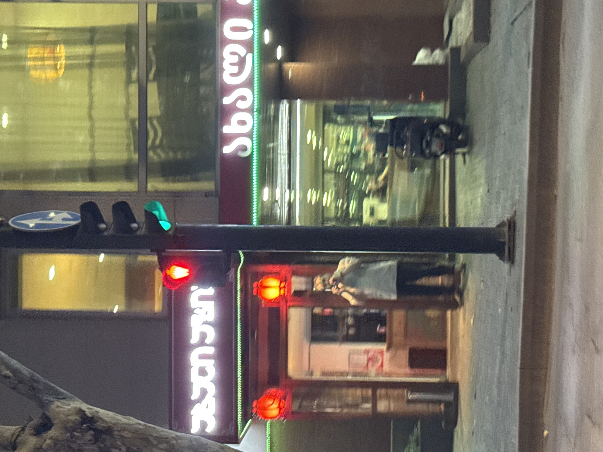 The image captures a street-level view at night in Tbilisi, Georgia, likely in an urban commercial area. A traffic light is prominently featured on the right side of the frame, with the red light illuminated. To the left of the traffic light, a building with a neon sign in Georgian script is visible. The sign glows white and appears to be in an alphabet with curved characters. Below the neon sign, there is an entrance to a establishment, marked by two red lanterns. A person, viewed from the chest up, is standing near the entrance, holding a phone up to their face, seemingly taking a picture or video. They are wearing a light-colored shirt and dark pants. In the background, through the windows of another establishment, blurry interior lights and reflections are discernible. A parked scooter is visible in front of this establishment. The sidewalk is paved with interlocking stones. A large tree trunk is partially visible on the far left of the image. The overall lighting suggests a nighttime scene with artificial illumination from signs, lights, and a traffic signal.