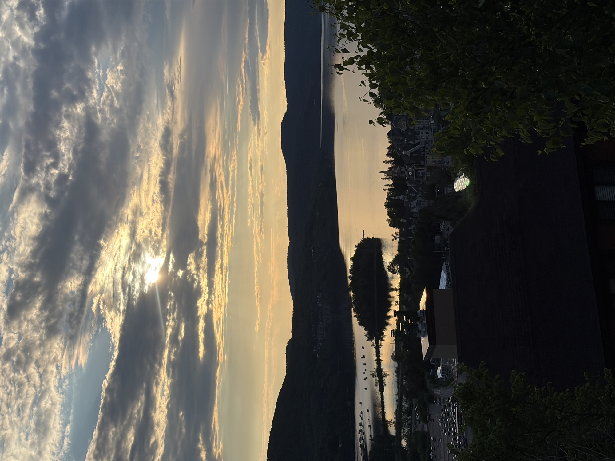 This is an aerial, high-angle view of a lake and surrounding landscape in Mont-Tremblant, Quebec, Canada, during sunset or sunrise. The sky is filled with dramatic clouds, with the sun partially obscured, casting a warm, golden light across the scene. A calm lake reflects the colors of the sky, with several small islands dotting its surface. On the far shore, dark, silhouetted mountains rise, with a cluster of buildings nestled at their base. In the foreground, the roofs of buildings and lush green foliage are visible. The image captures a serene and picturesque view of the natural beauty and the charming architecture of Mont-Tremblant.